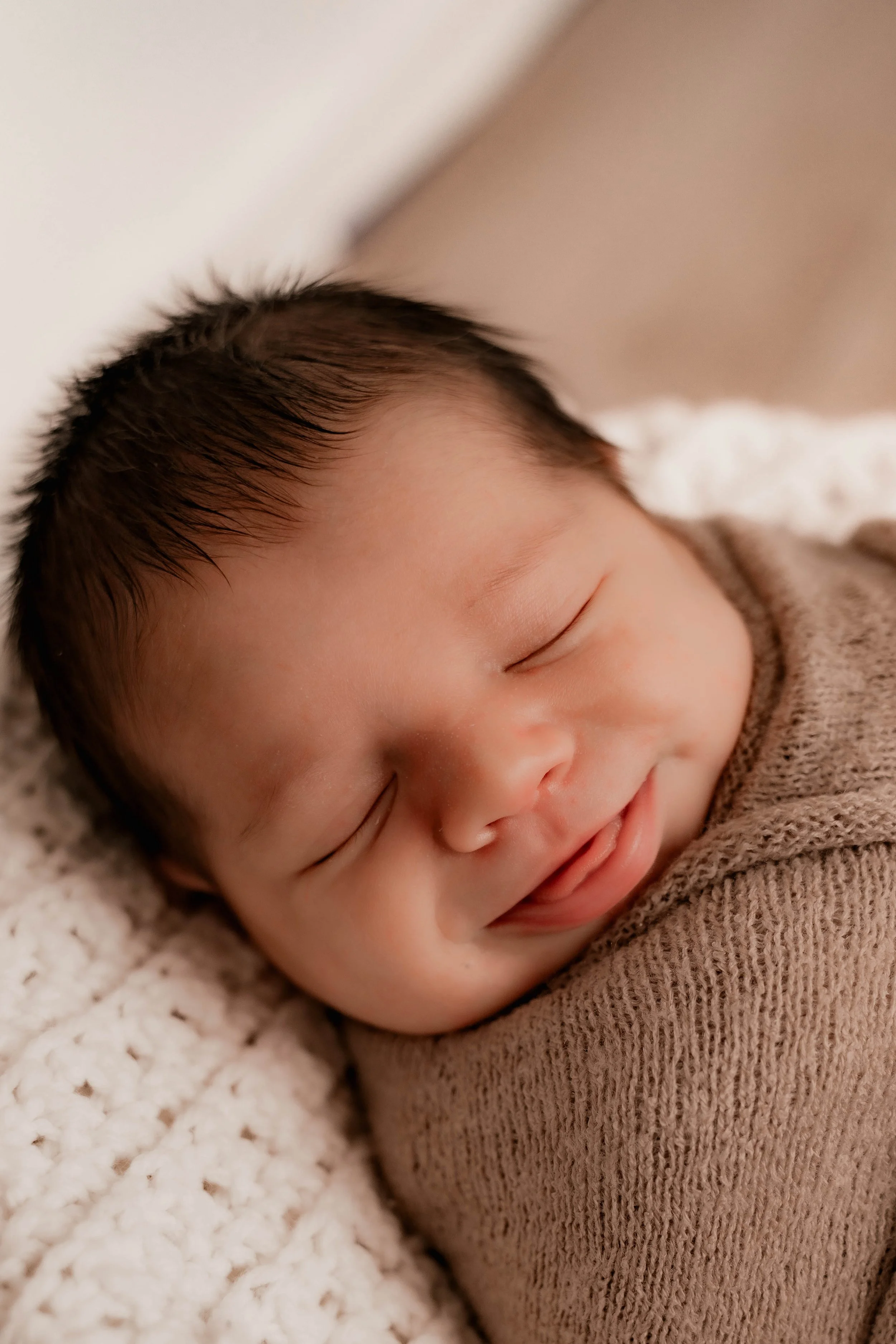 Close-up of a smiling baby sleeping on a soft, textured blanket, wearing a brown knitted outfit.