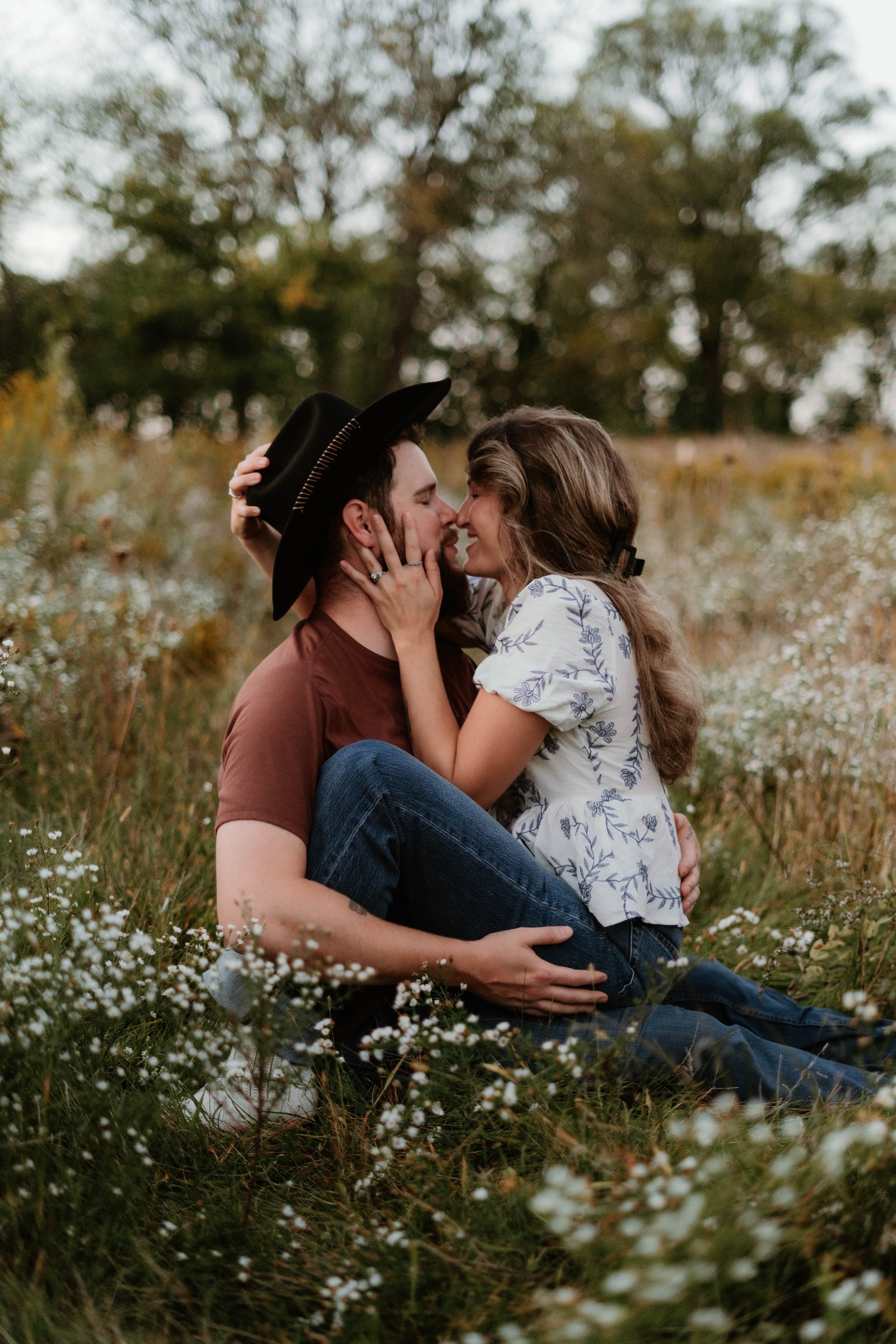 A couple sitting in a field of small white flowers, close together, about to kiss. The man is wearing a brown shirt and a black cowboy hat, and the woman is wearing a white blouse with blue floral patterns. She has her hands on his face, and he is ho