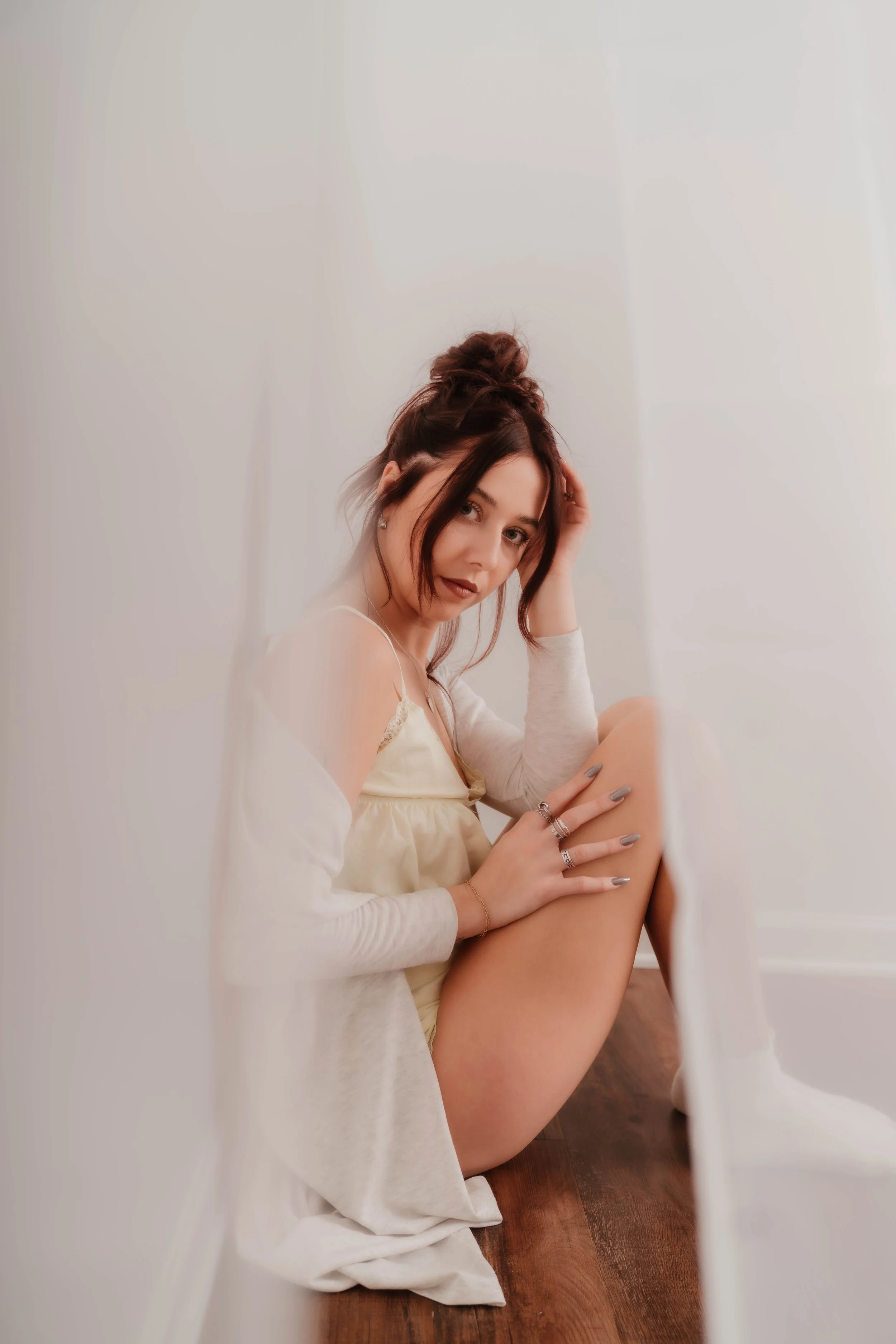 A young woman sitting on a hardwood floor, gazing at the camera, wearing a cream-colored slip dress and a light cardigan, with her hair in a messy bun and hand resting on her head, surrounded by soft white walls.
