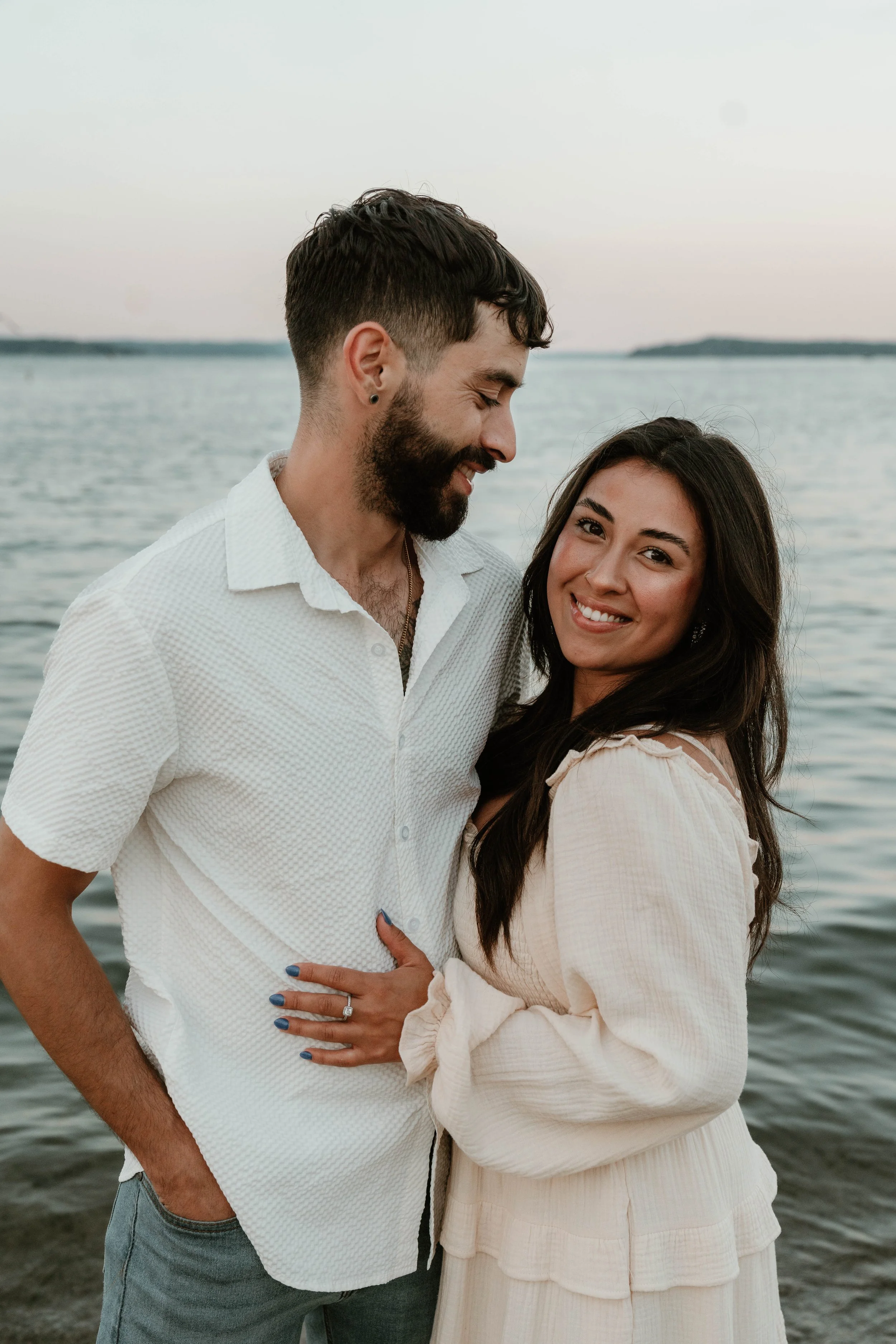 A couple standing close together at the water's edge, with the man smiling and looking down at the woman who is smiling at the camera, during sunset or early evening.