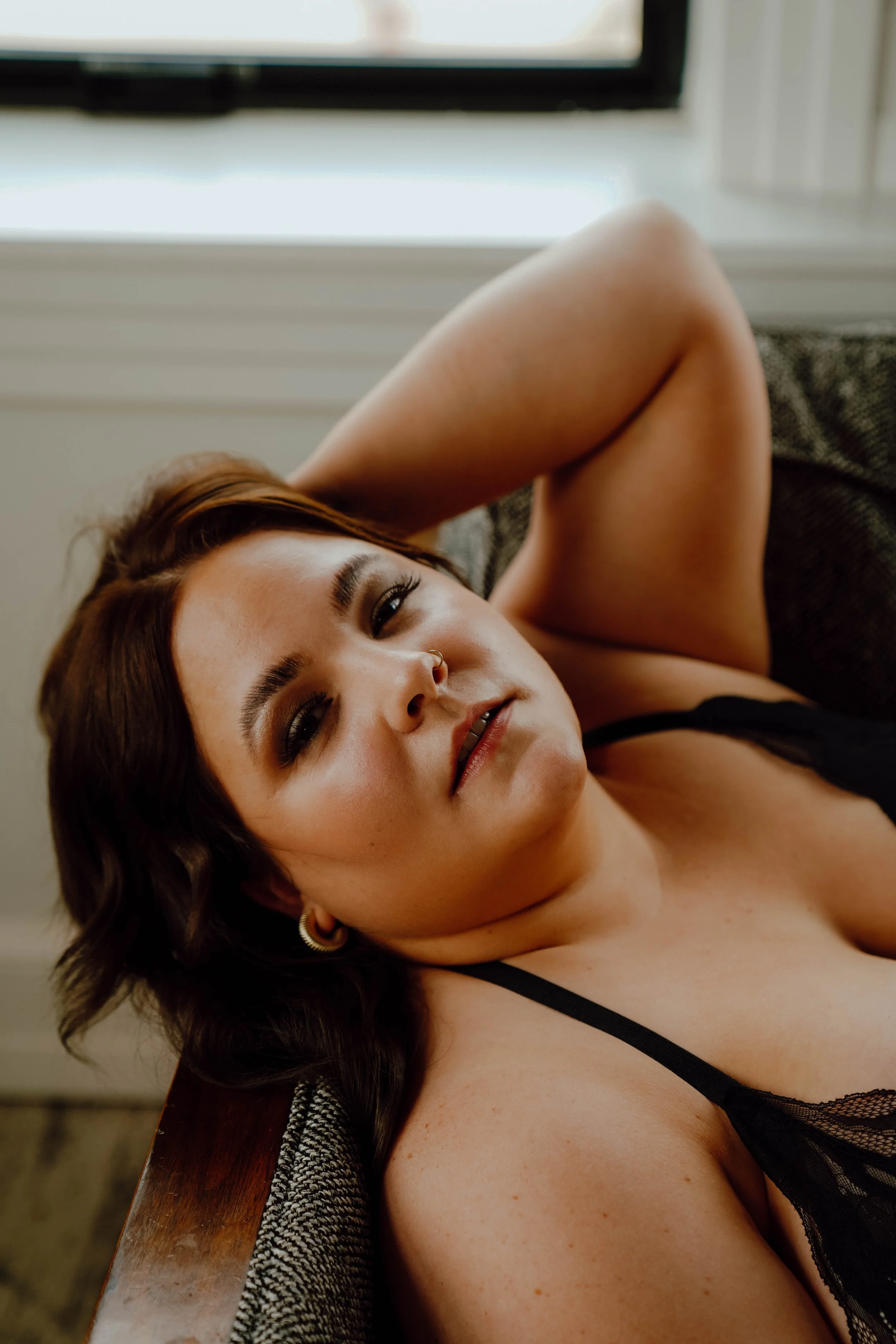 A woman with dark hair and makeup lying on a couch, wearing a black lacy top, with one arm behind her head and a window in the background.