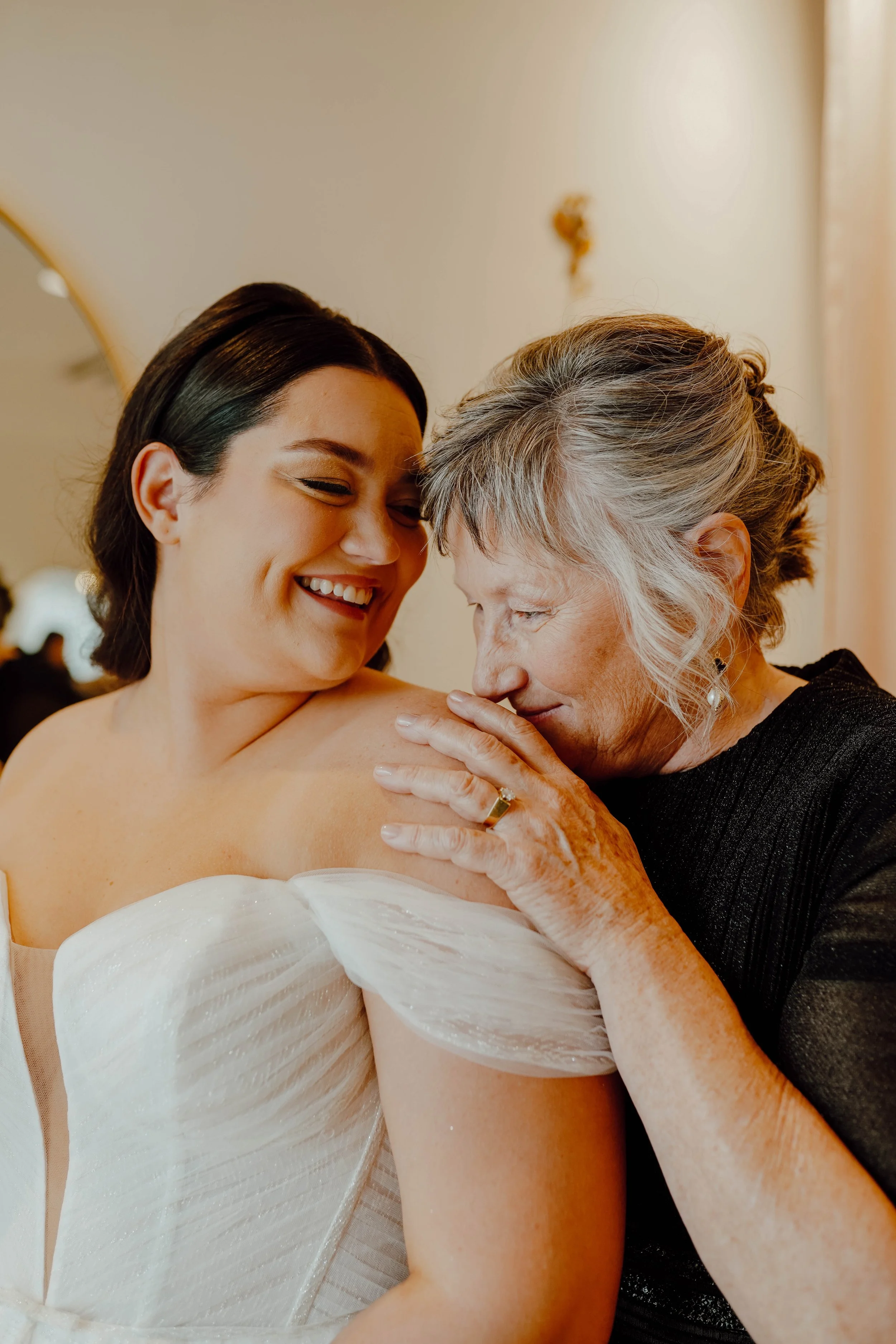 A young woman in a wedding dress with dark hair smiling and an older woman with gray hair embracing her gently, touching her shoulder.