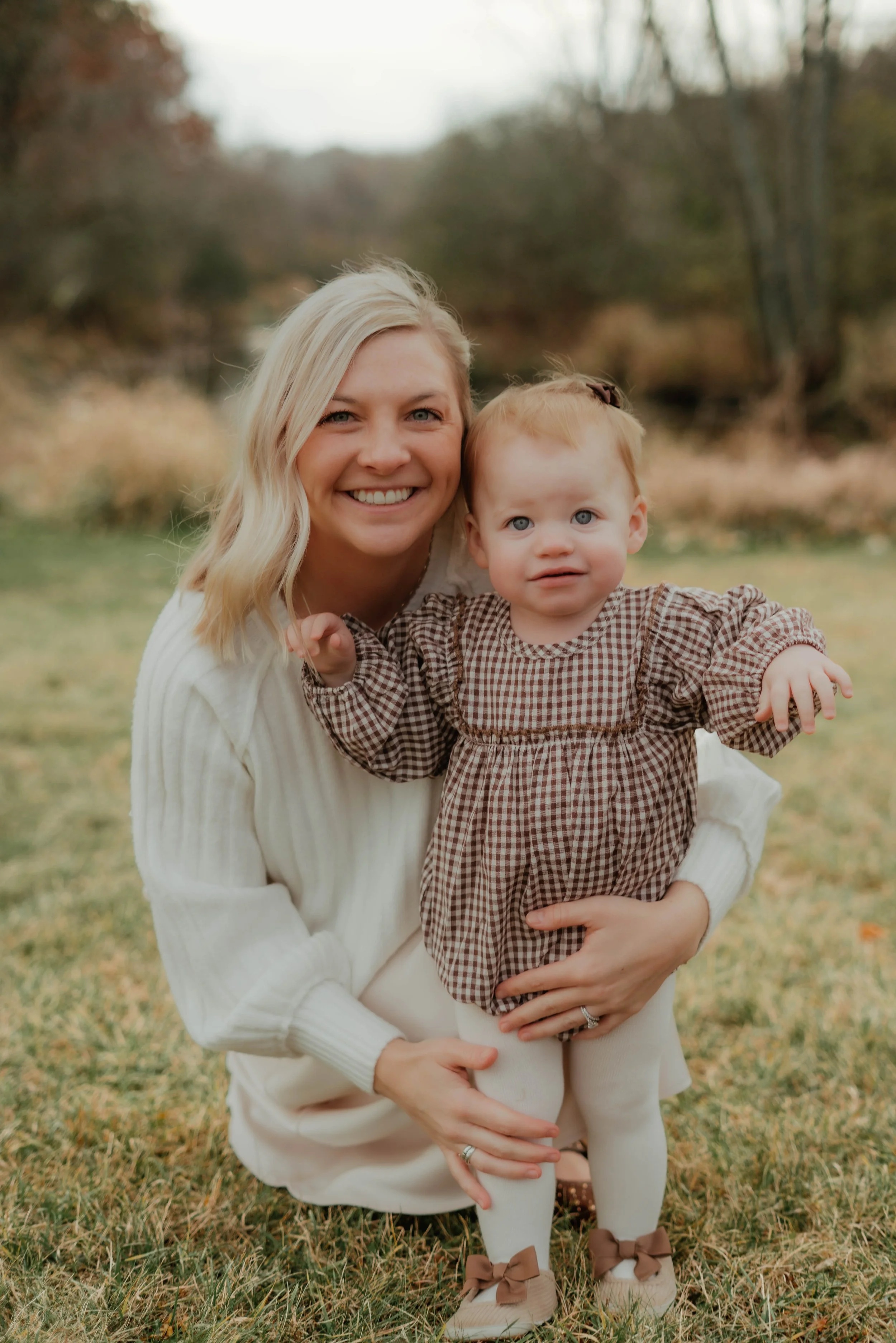 A smiling blonde woman kneeling outdoors on grass, holding a young girl with red hair and blue eyes, both dressed in brown checkered outfits, in a park setting during fall with trees and overcast sky in the background.