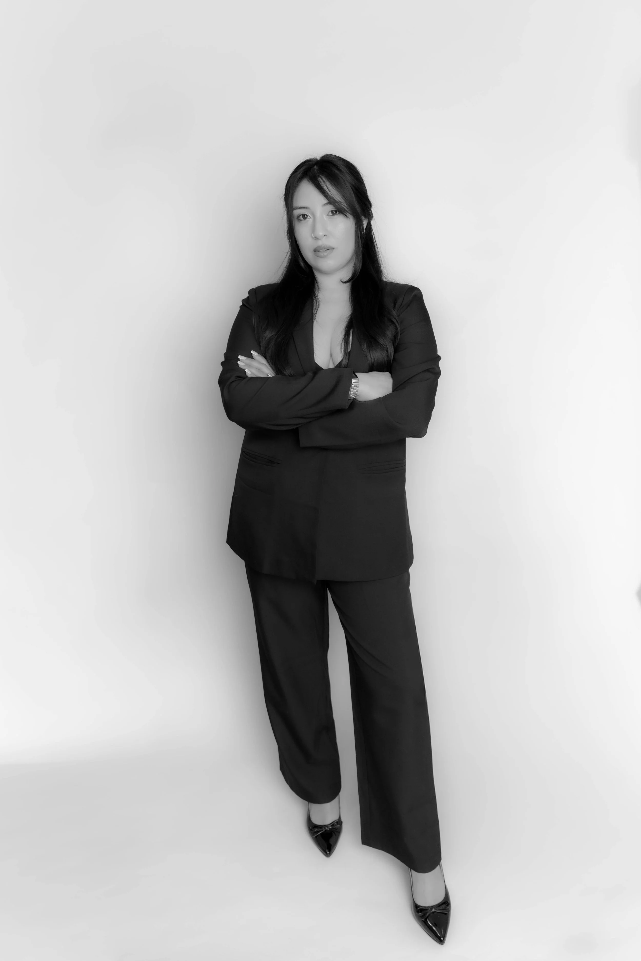A woman with dark hair and light skin standing against a plain white background, wearing a black suit with her arms crossed.