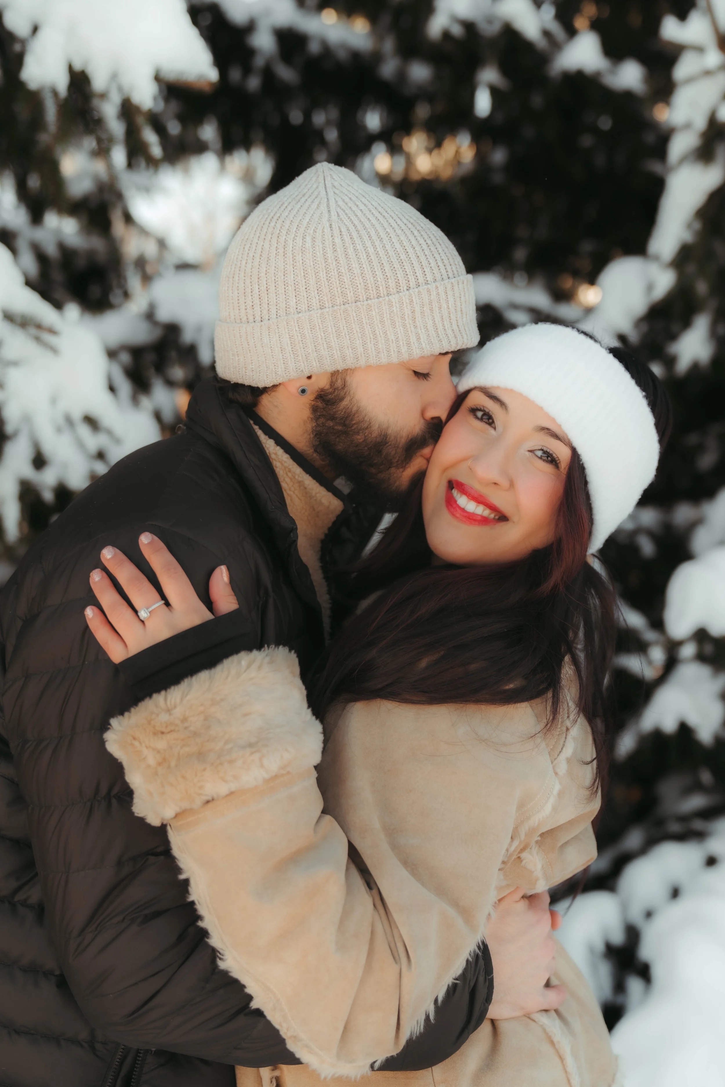 A couple embracing outdoors in a snowy setting, with the man kissing the woman on the cheek. Both are dressed warmly in winter clothing, with white beanies and a winter coat.