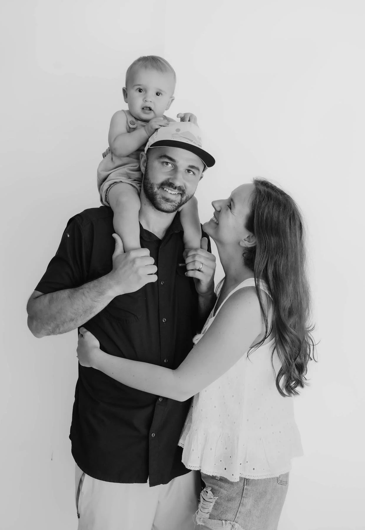 Happy family of three with a man carrying a toddler on his shoulders and a woman embracing him, all smiling and looking at each other, in a studio with a plain background.
