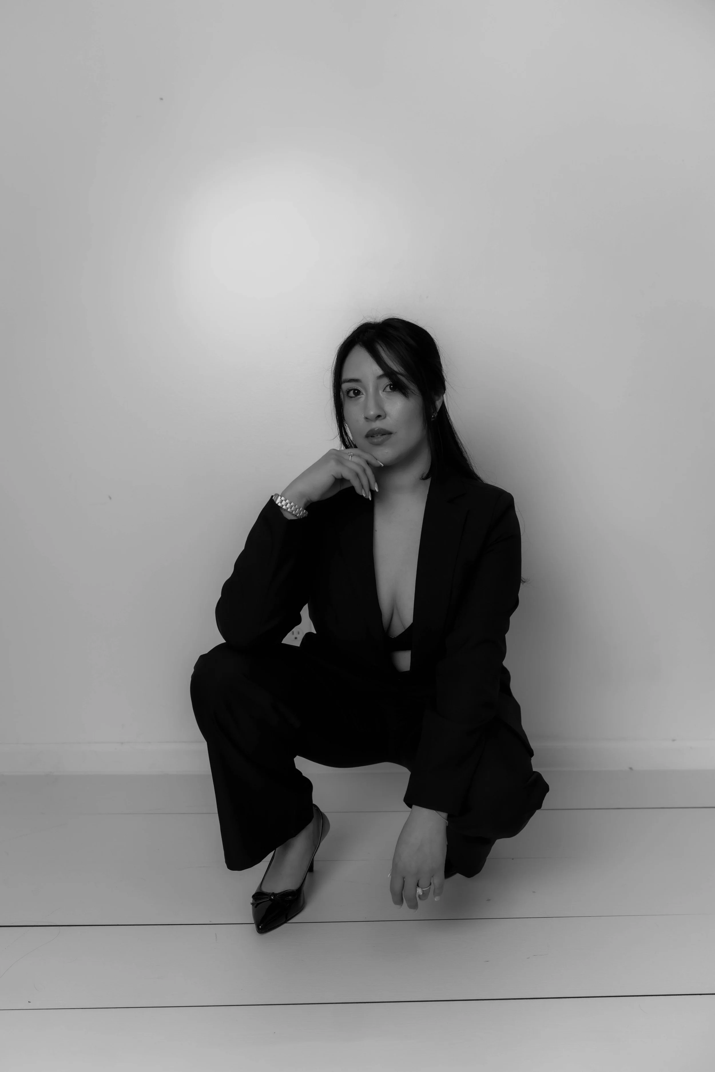 A woman squatting on the floor against a plain wall, wearing a dark blazer and pants, with high heels, and resting her chin on her hand.
