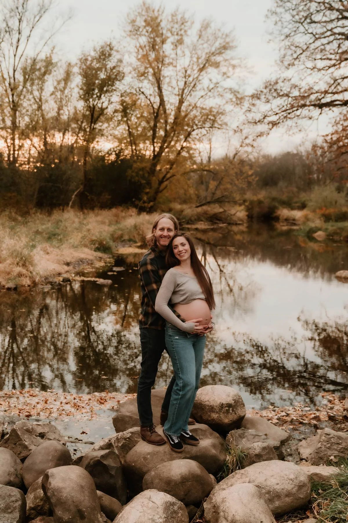 A pregnant woman and a man are standing on rocks near a river with trees and a sunset in the background.