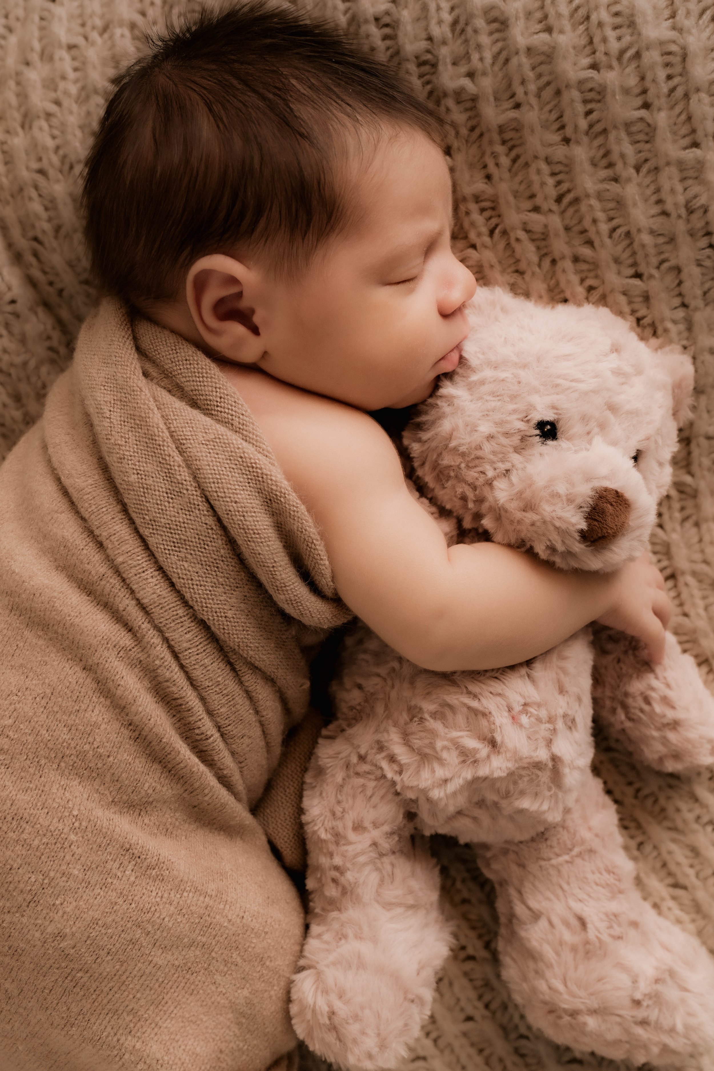A sleeping young child hugging a plush teddy bear while laying on a textured, brown knitted blanket.