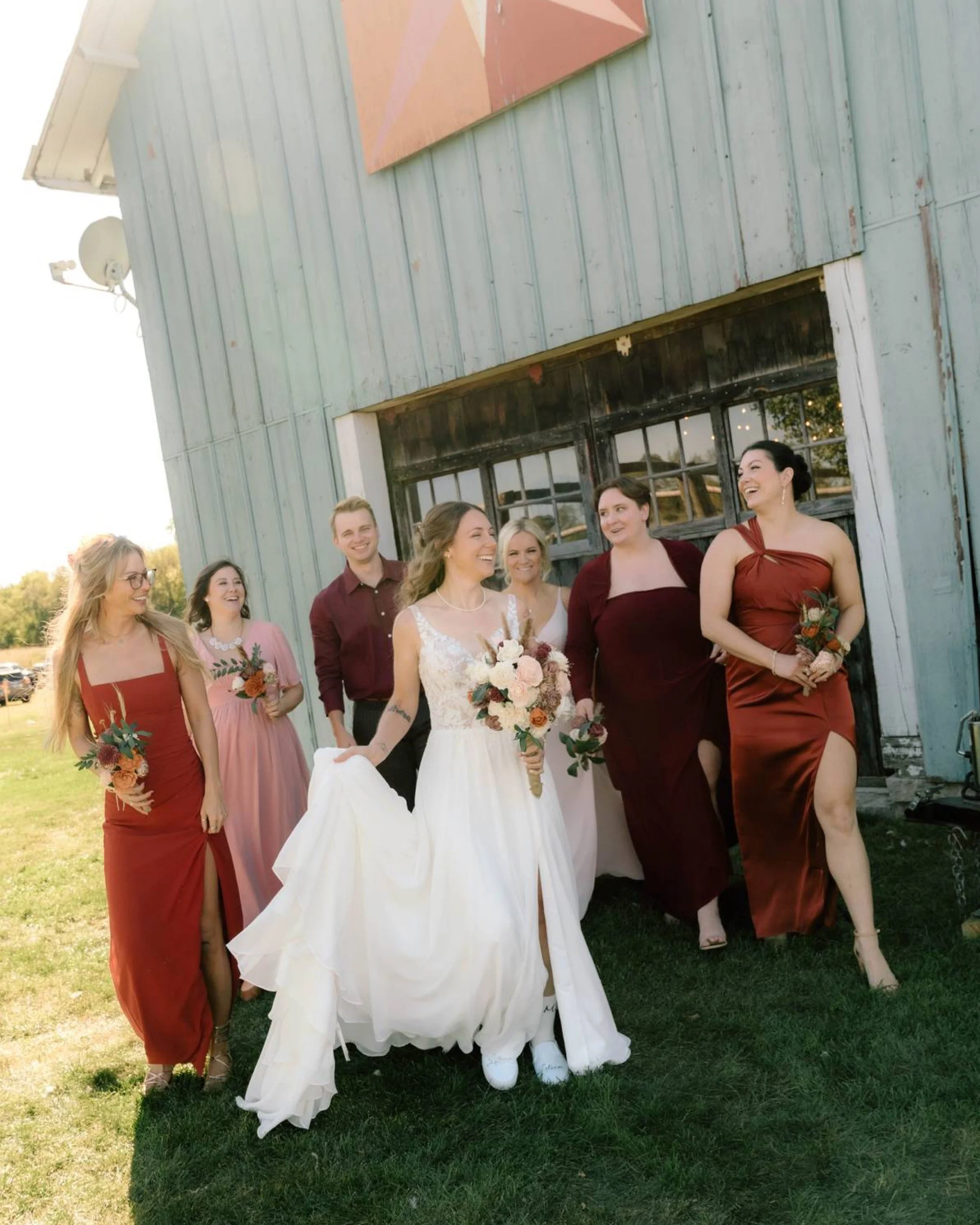 Group of women, including a bride in a white wedding dress, and bridesmaids in burgundy and pink dresses, walking and smiling outdoors in front of a rustic building with distressed blue wooden siding.