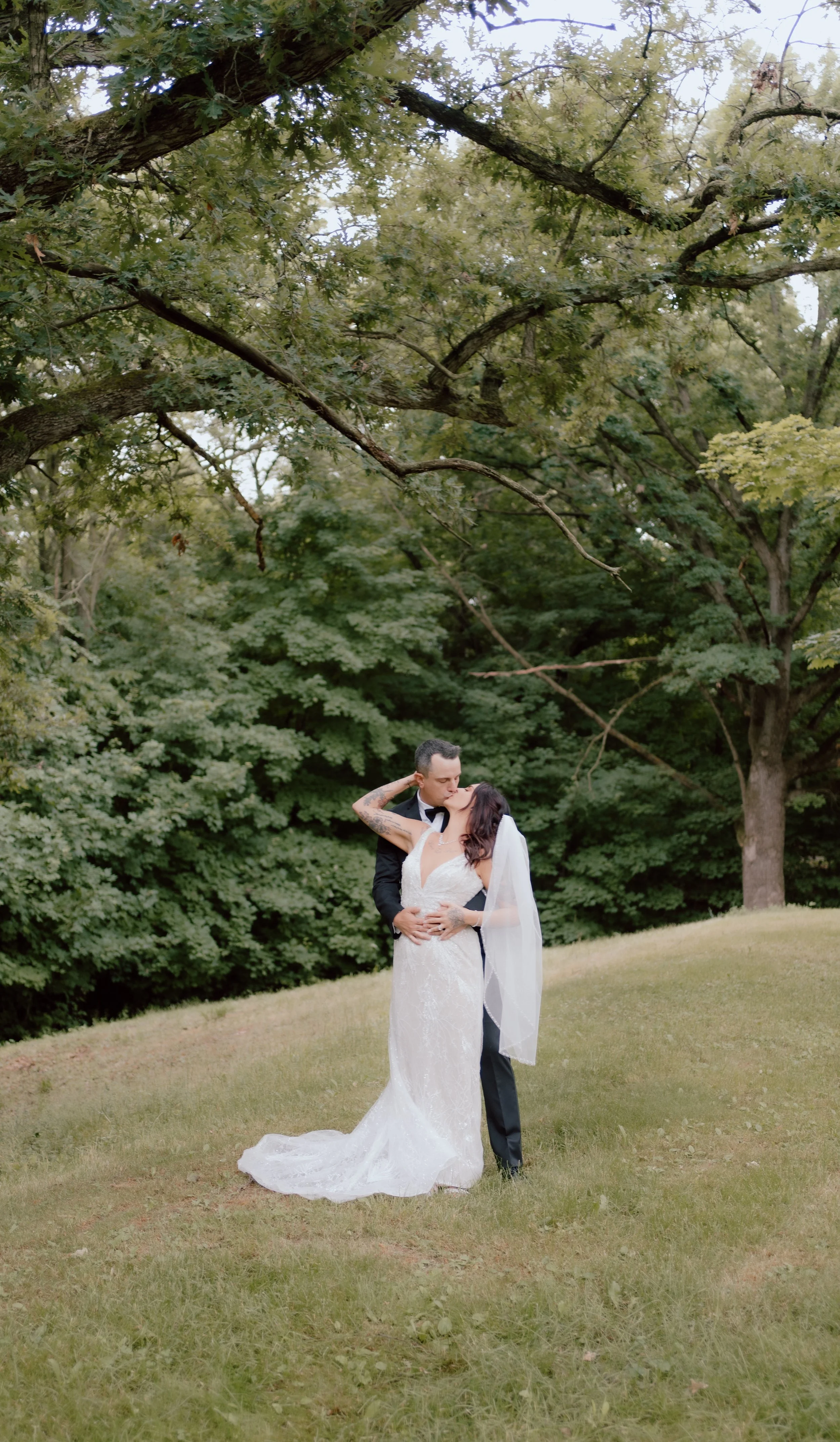 A bride and groom kissing outdoors on a grassy area surrounded by trees during their wedding.