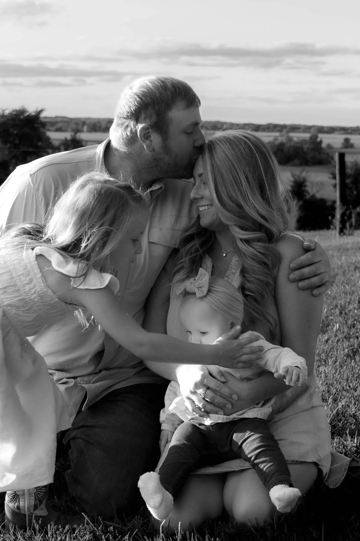 A black and white photo of a family outdoors. The man is kissing the woman's forehead while she smiles. Two young girls are interacting with a toddler girl who is sitting on the woman's lap, reaching out to touch the toddler's arm. The background sho