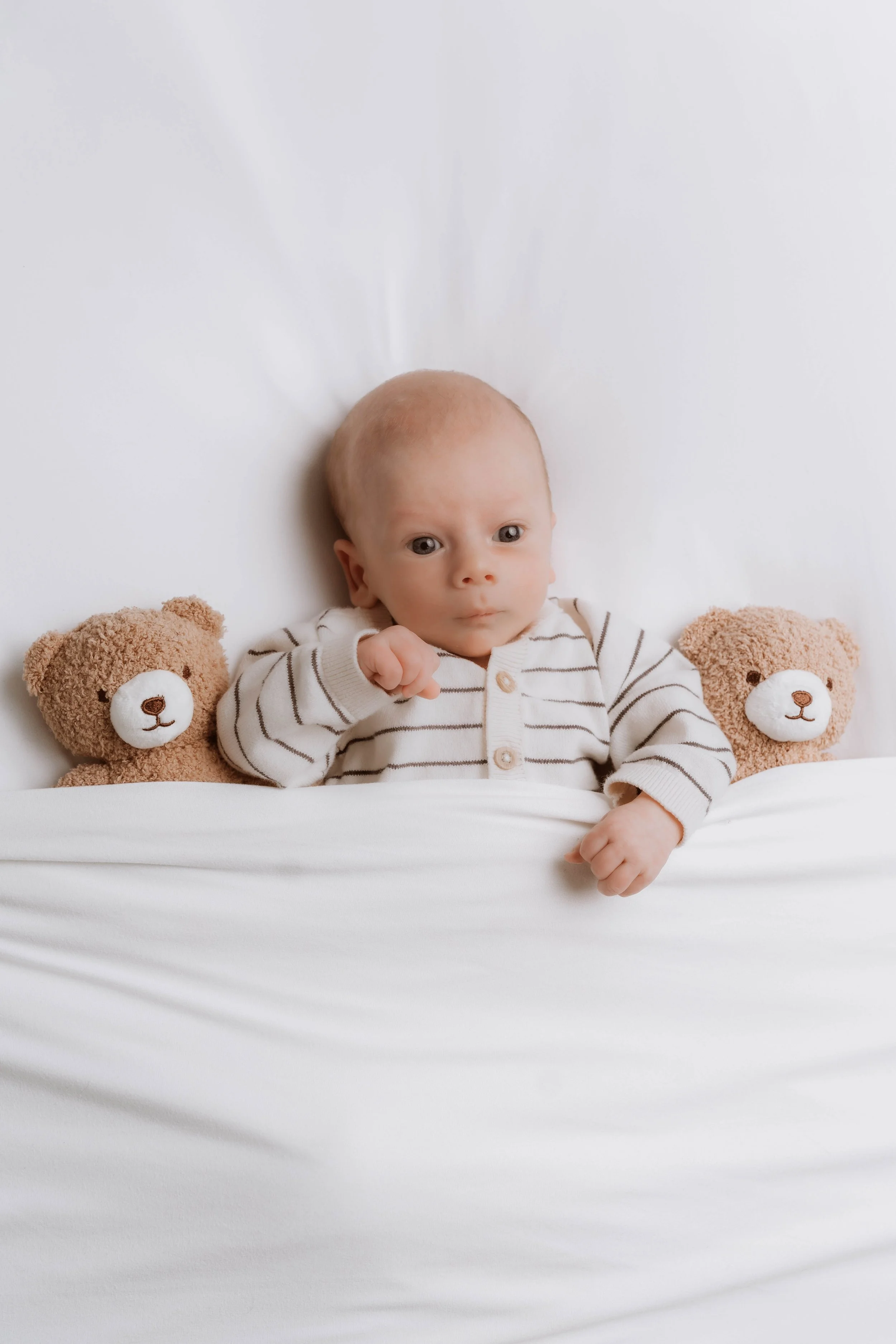 Cute baby lying in bed between two teddy bears, wearing a striped shirt.