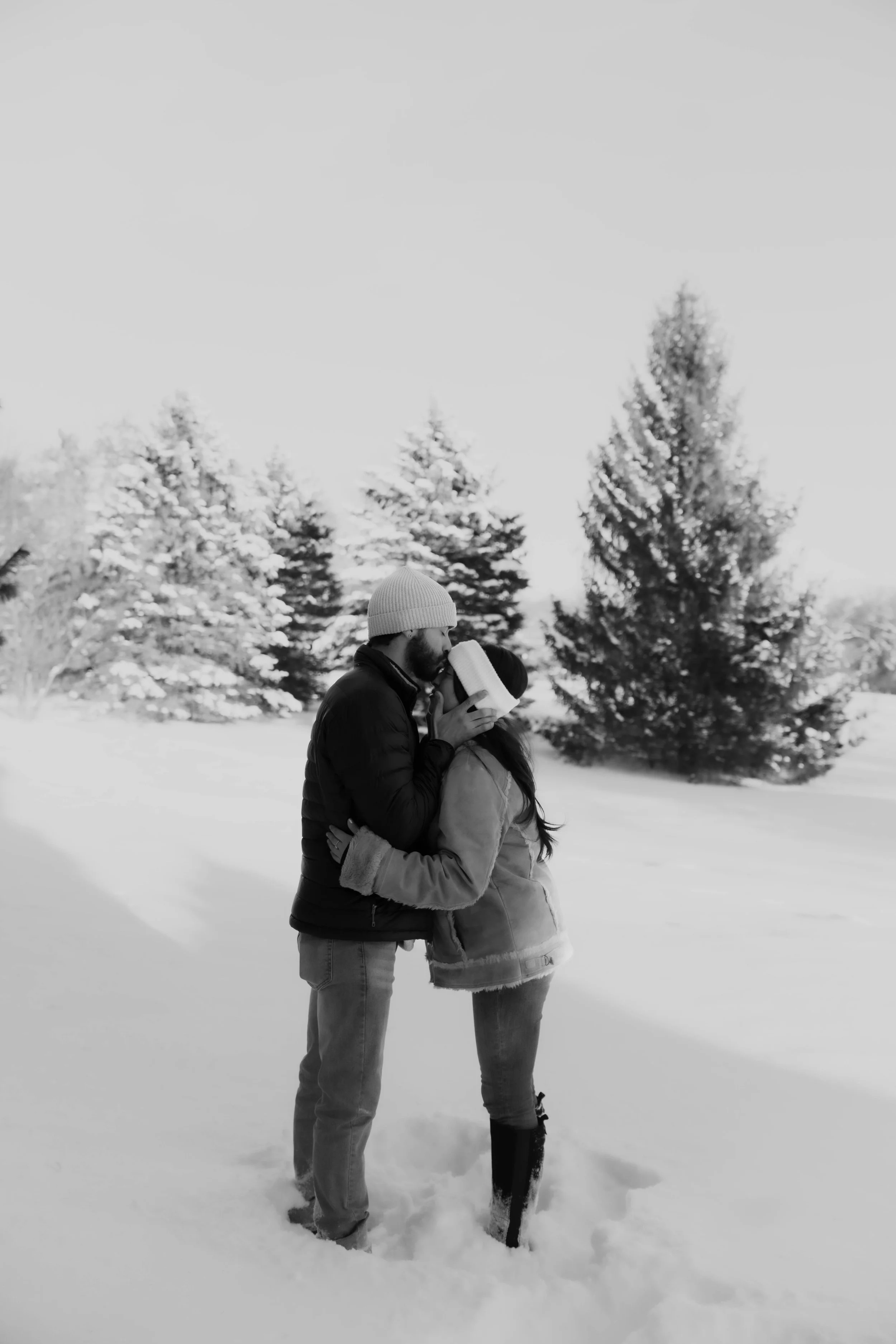 A couple in winter clothing sharing a kiss in the snow during daytime, with snow-covered trees in the background.