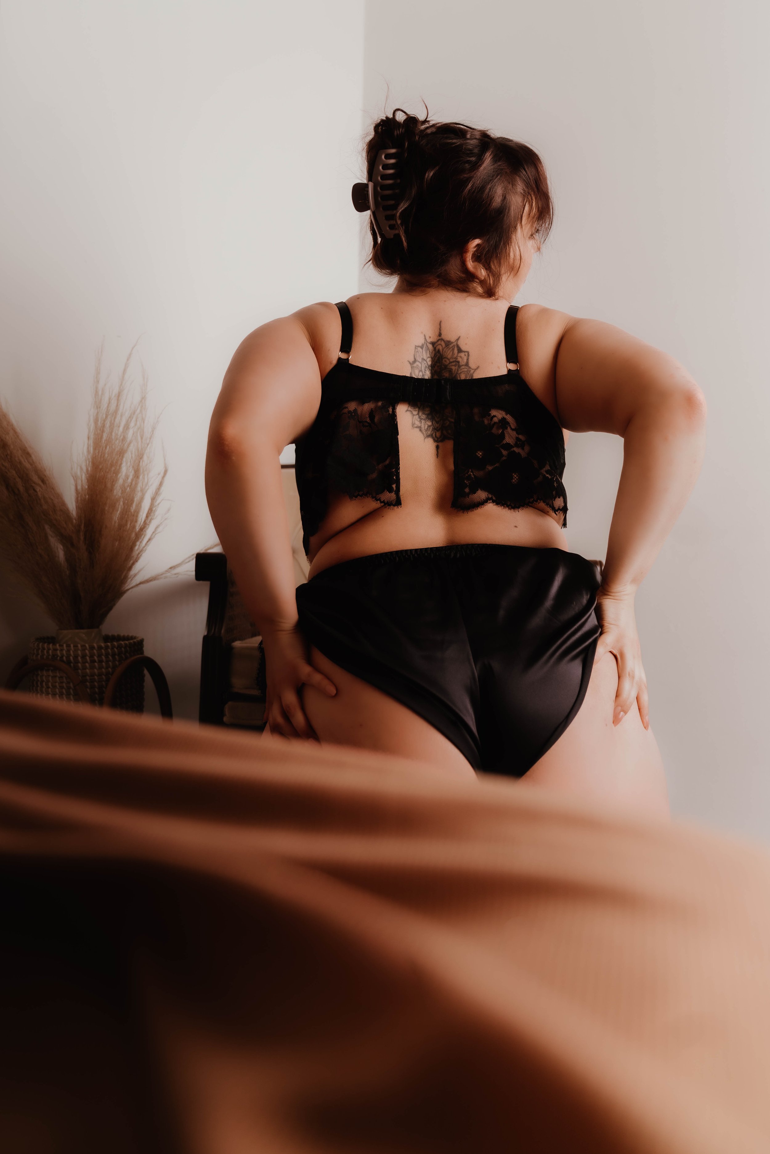 A woman with dark hair in a hair clip, wearing a black lace top and black satin shorts, standing with her back to the camera, touching her thigh, in a softly lit room with neutral-colored walls and a decorative vase with dried plants.