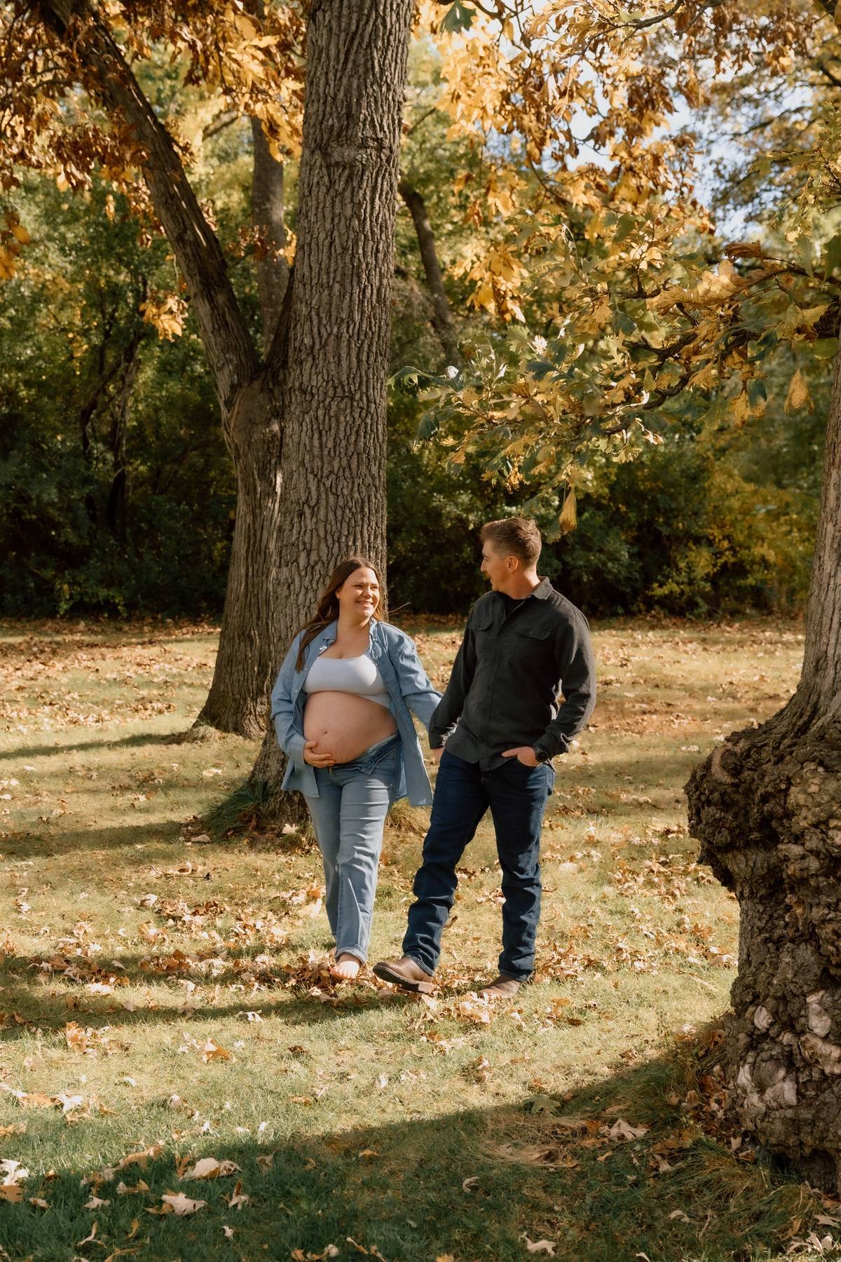 A pregnant woman and a man walking in a park during autumn. The woman is holding her belly and smiling at the man, both are dressed casually. The scene features trees with autumn leaves and a grassy area.