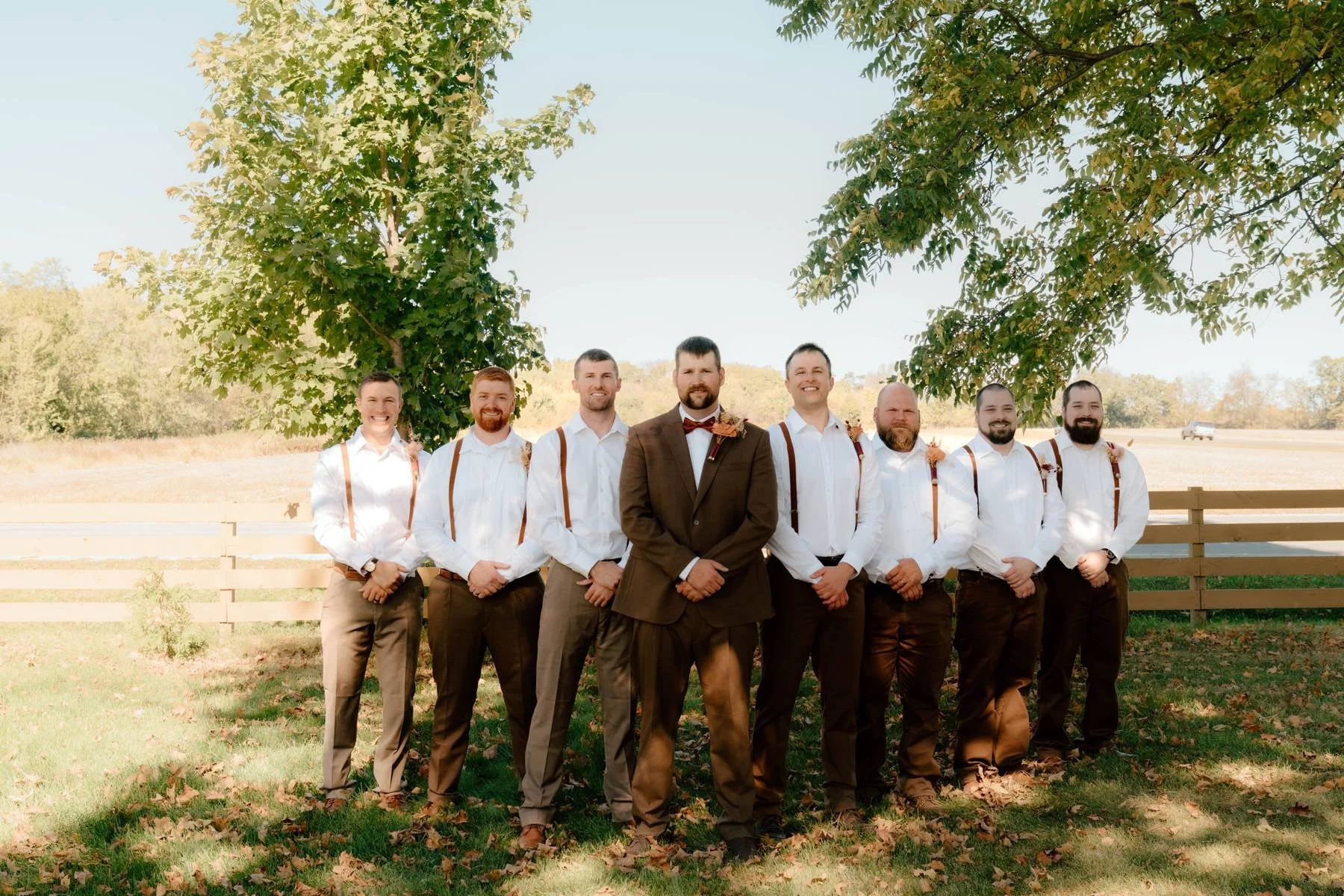 A group of nine men in formal attire standing outdoors under a large tree on a sunny day. The man in the center is wearing a brown suit and bow tie, while the others are dressed in white shirts, brown pants, and suspenders.