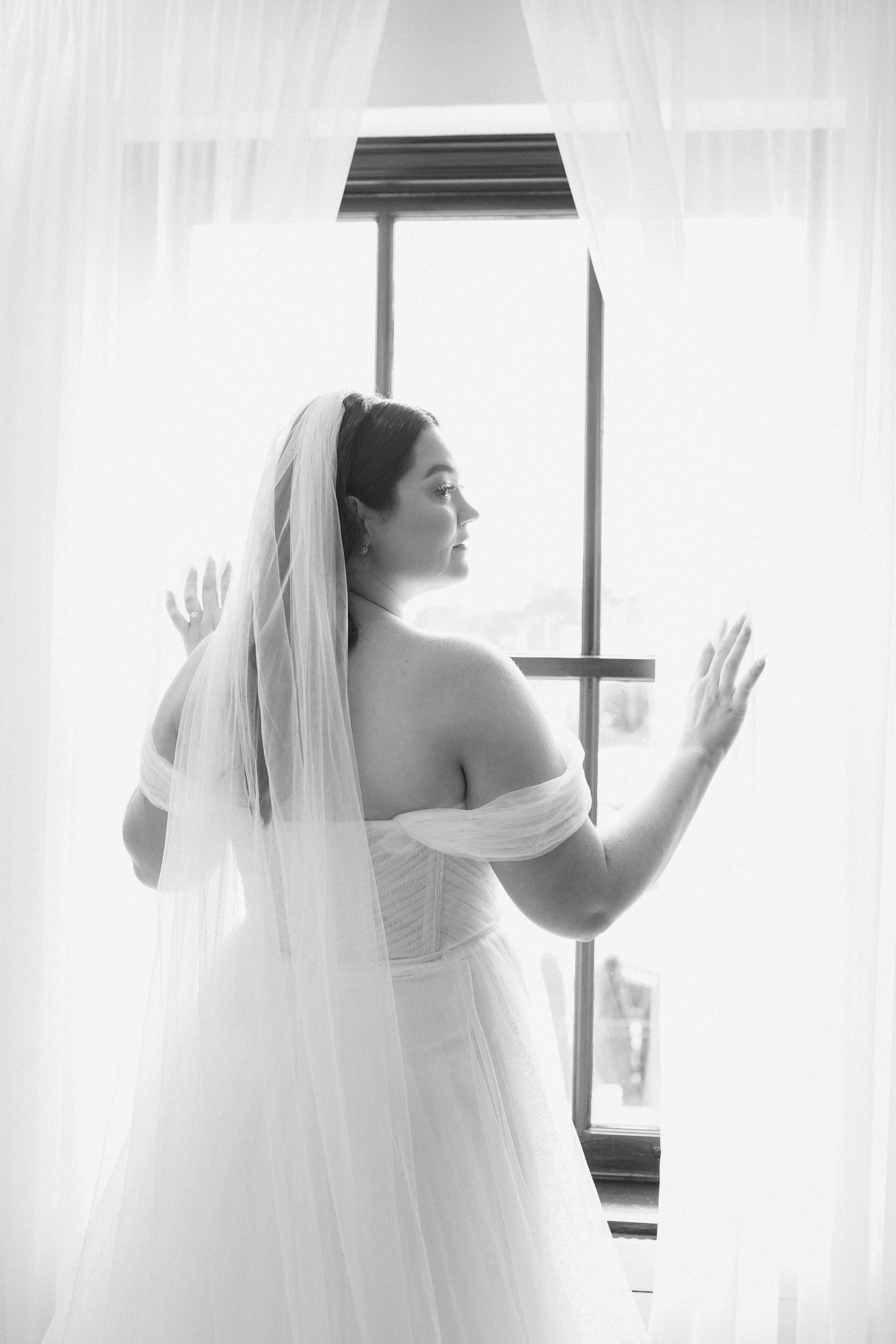 A woman in a wedding dress and veil standing by a window, looking outside, with her hands gently touching the curtains.