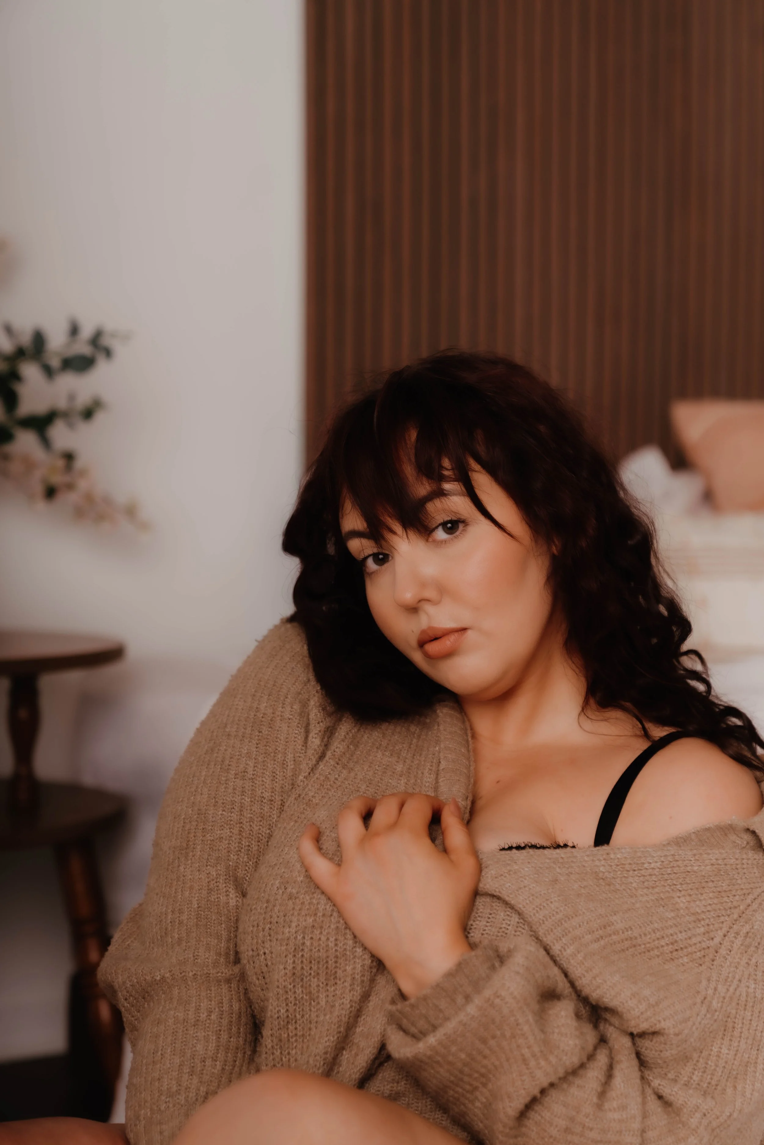 A young woman with dark, curly hair and light skin is sitting indoors, wearing a beige sweater and a black bra strap visible on her left shoulder. She is resting her head on her right hand and looking directly at the camera with a neutral expression.