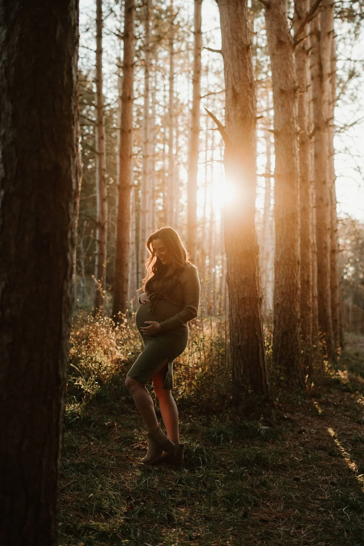 Pregnant woman standing outdoors in a forest during sunset, holding her belly and smiling.
