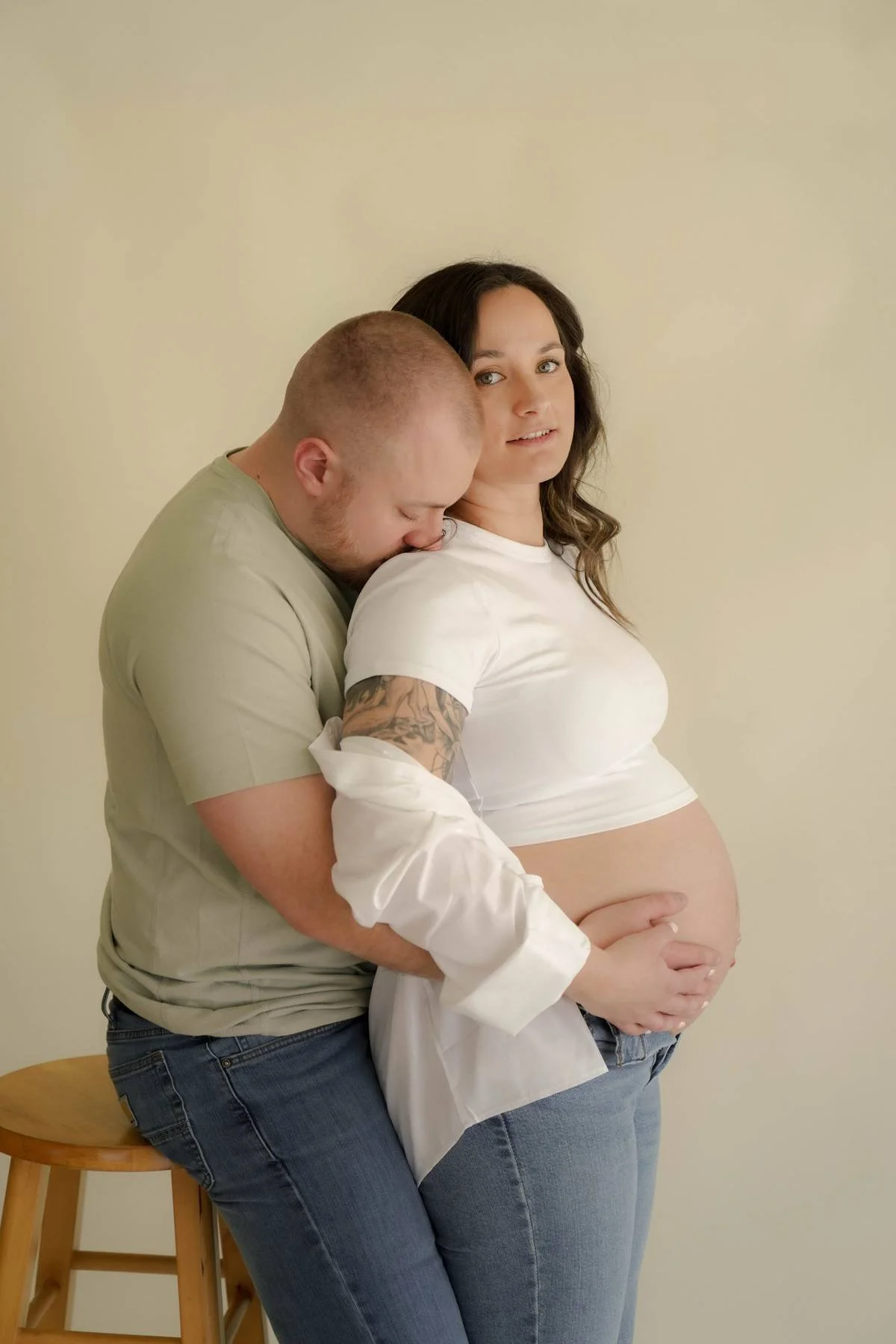 Pregnant woman with dark hair in a white t-shirt and jeans, holding her belly, with a man behind her, leaning on her shoulder with his head on her chest. They are standing against a plain light-colored wall. The man has short hair, a beard, and tatto