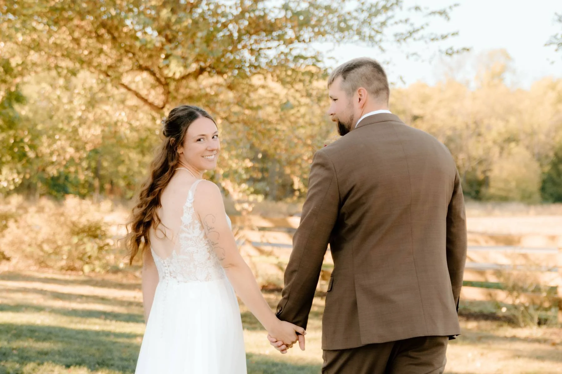 A bride and groom holding hands outdoors during their wedding, with autumn trees in the background.