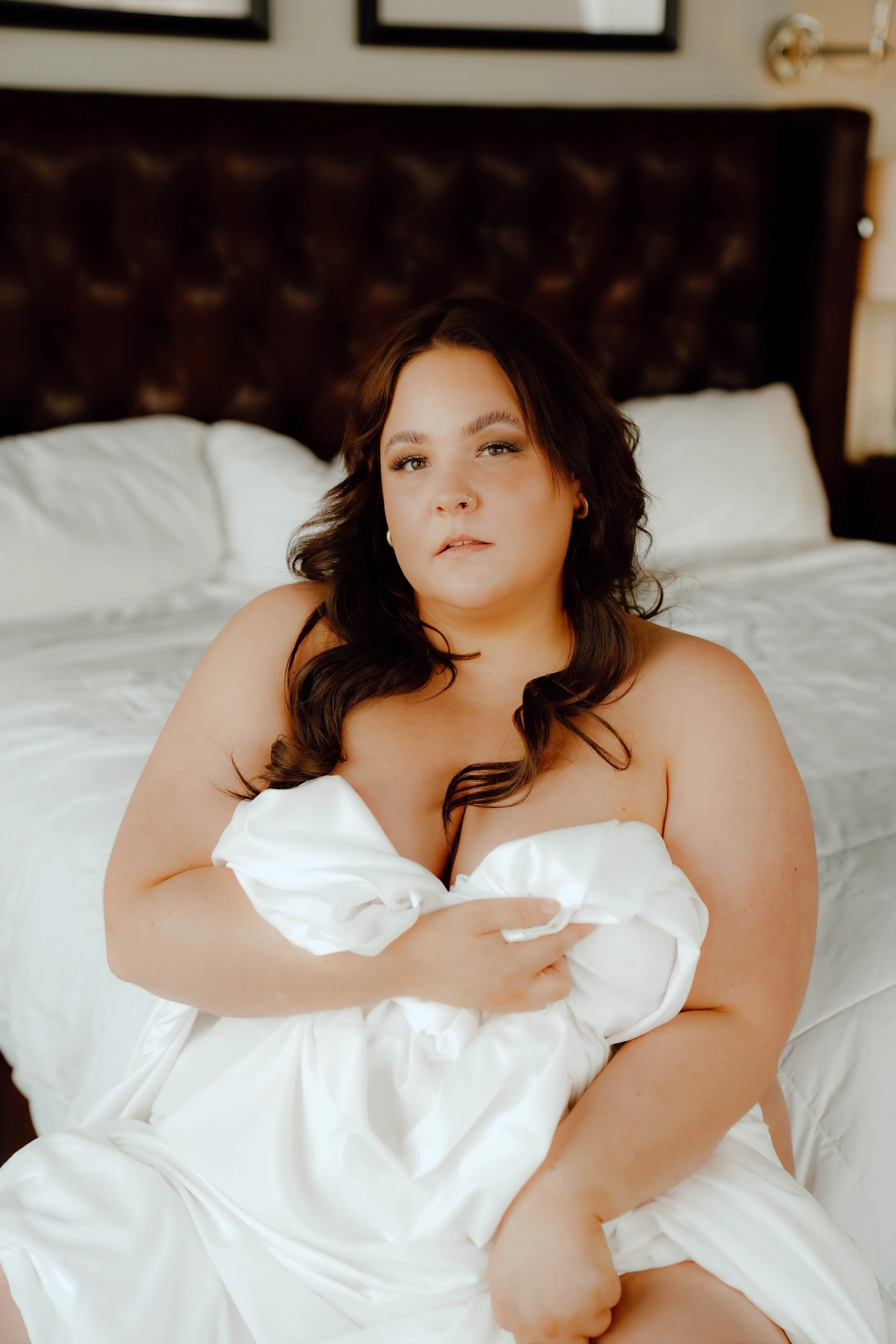 A woman with dark wavy hair sitting on a bed, holding a white bedsheet, with an upholstered headboard behind her in a bedroom.