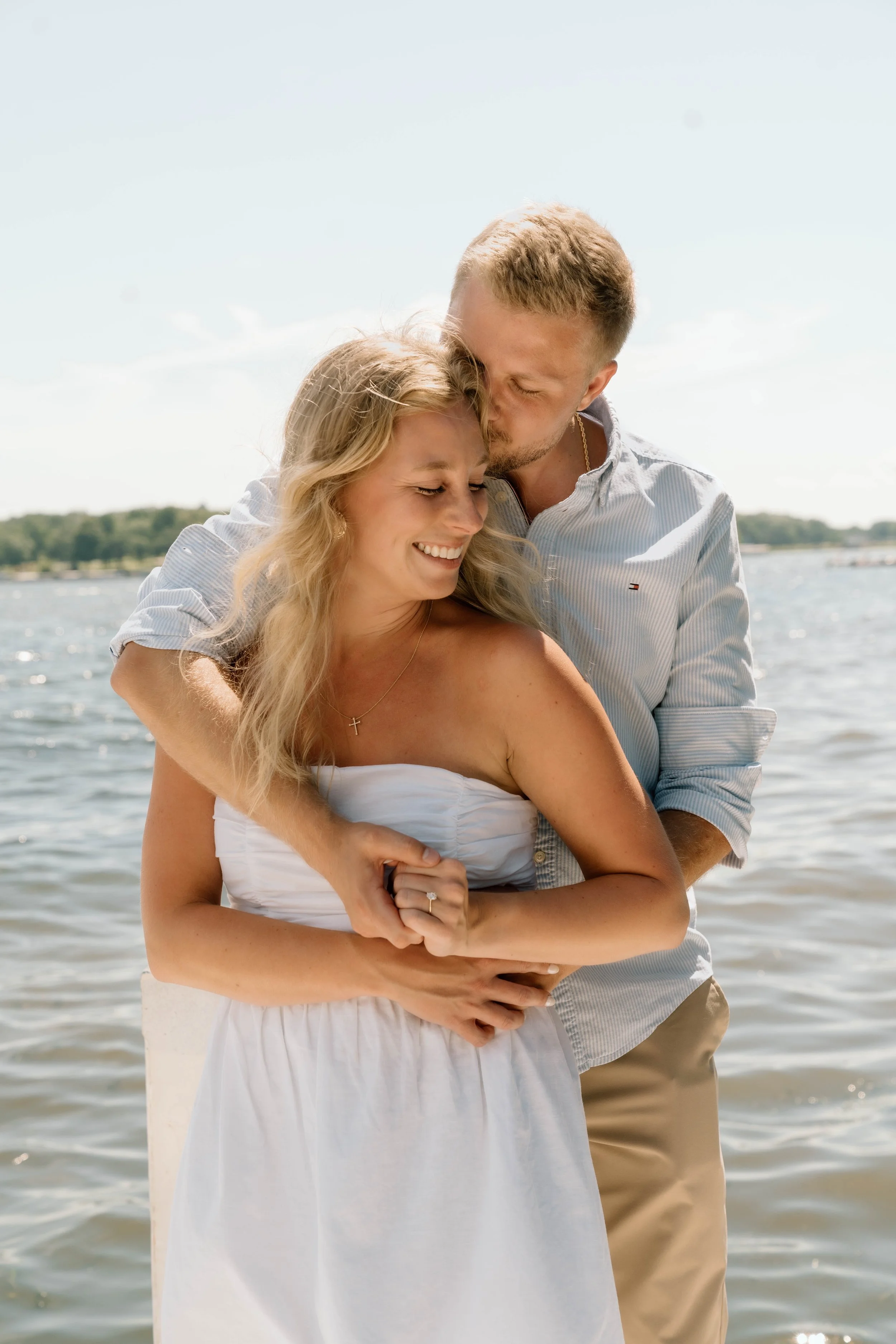 A smiling couple embracing near a body of water, with the man kissing the woman's forehead on a sunny day