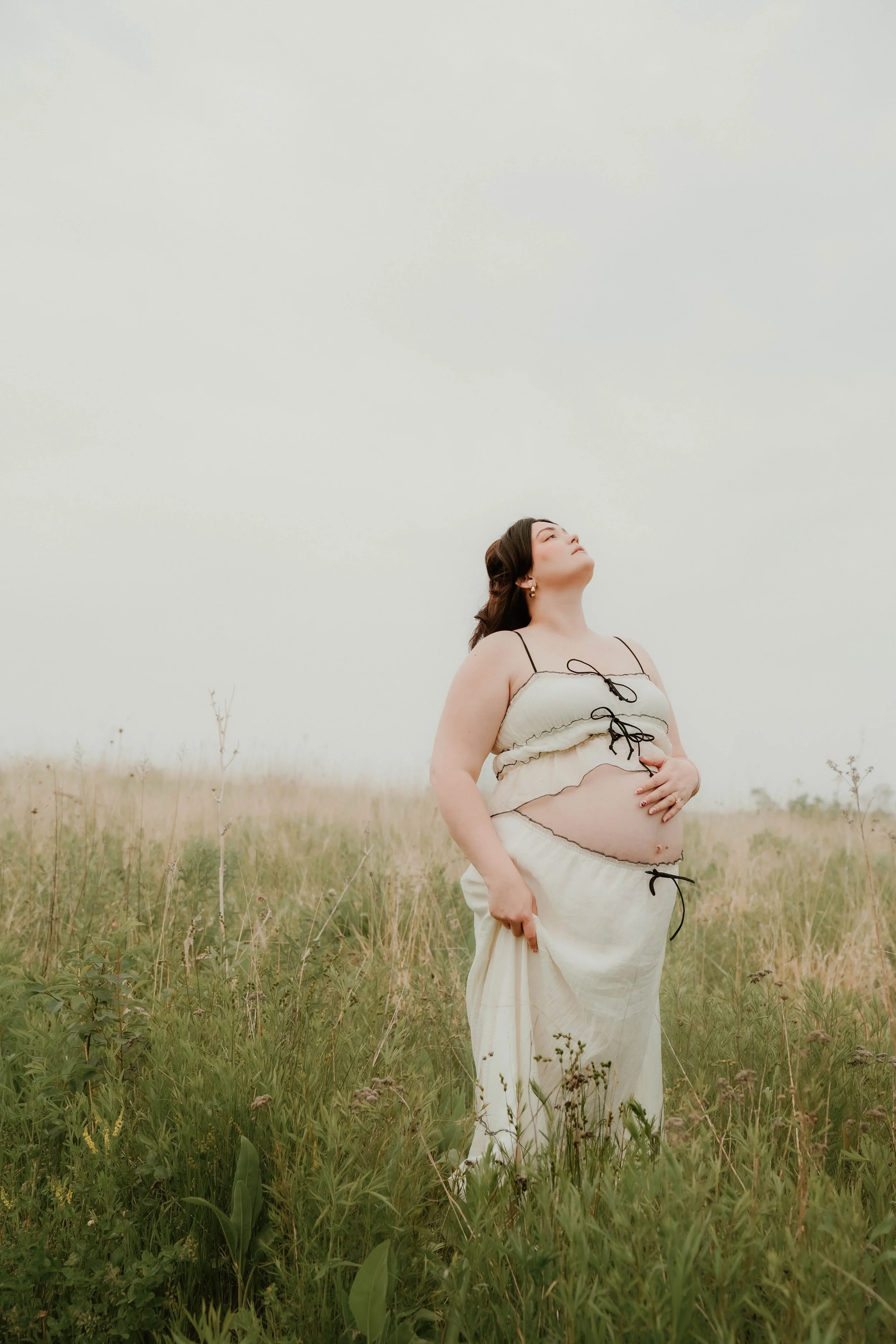 A woman standing in a grassy field with her eyes closed, wearing a cream-colored, layered dress with black tie details, holding up part of her dress with her right hand, and gently touching her stomach with her left hand.