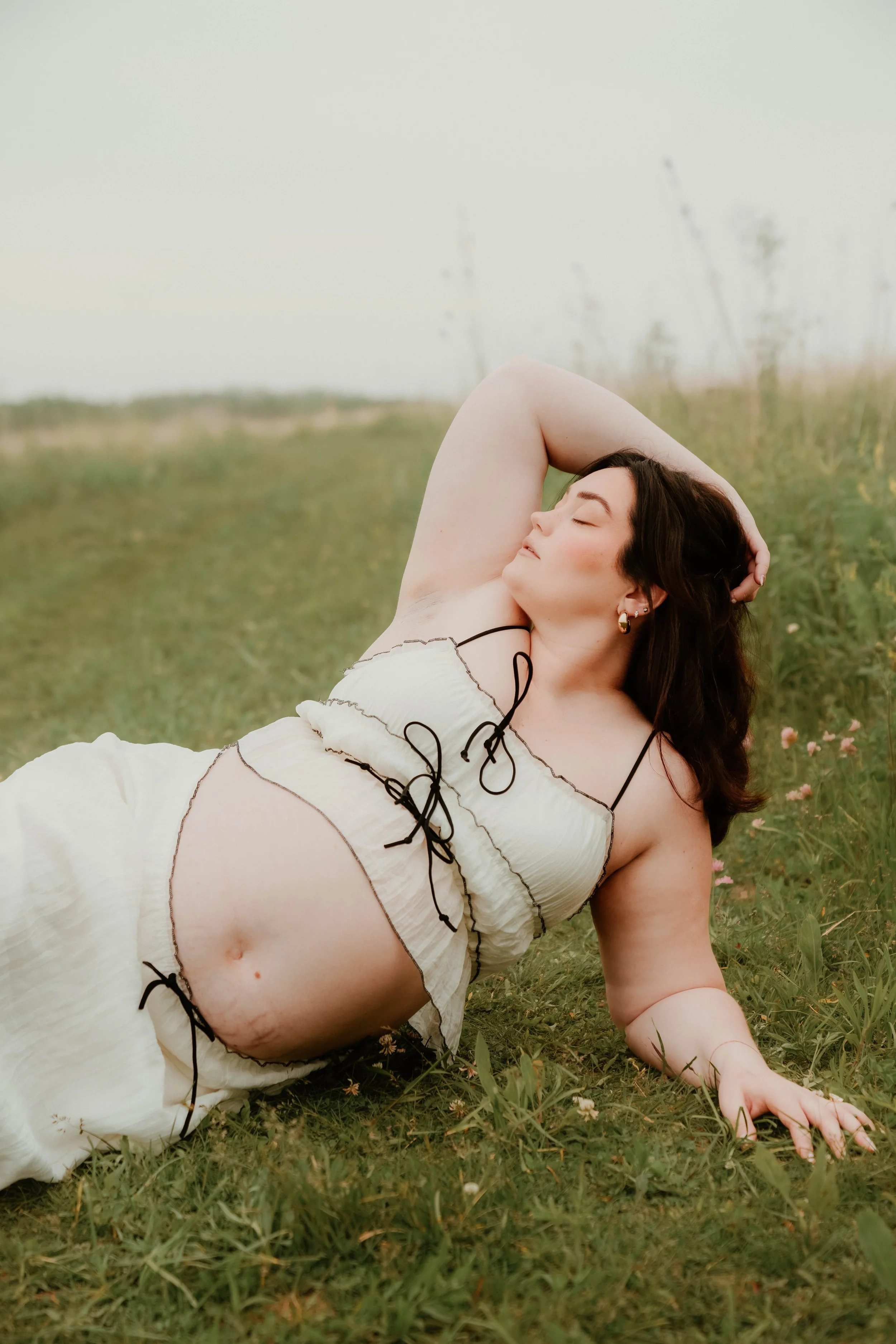 A woman with dark curly hair lying on the grass in a field, wearing a cream-colored dress with black strings, eyes closed, with one arm resting on her head.