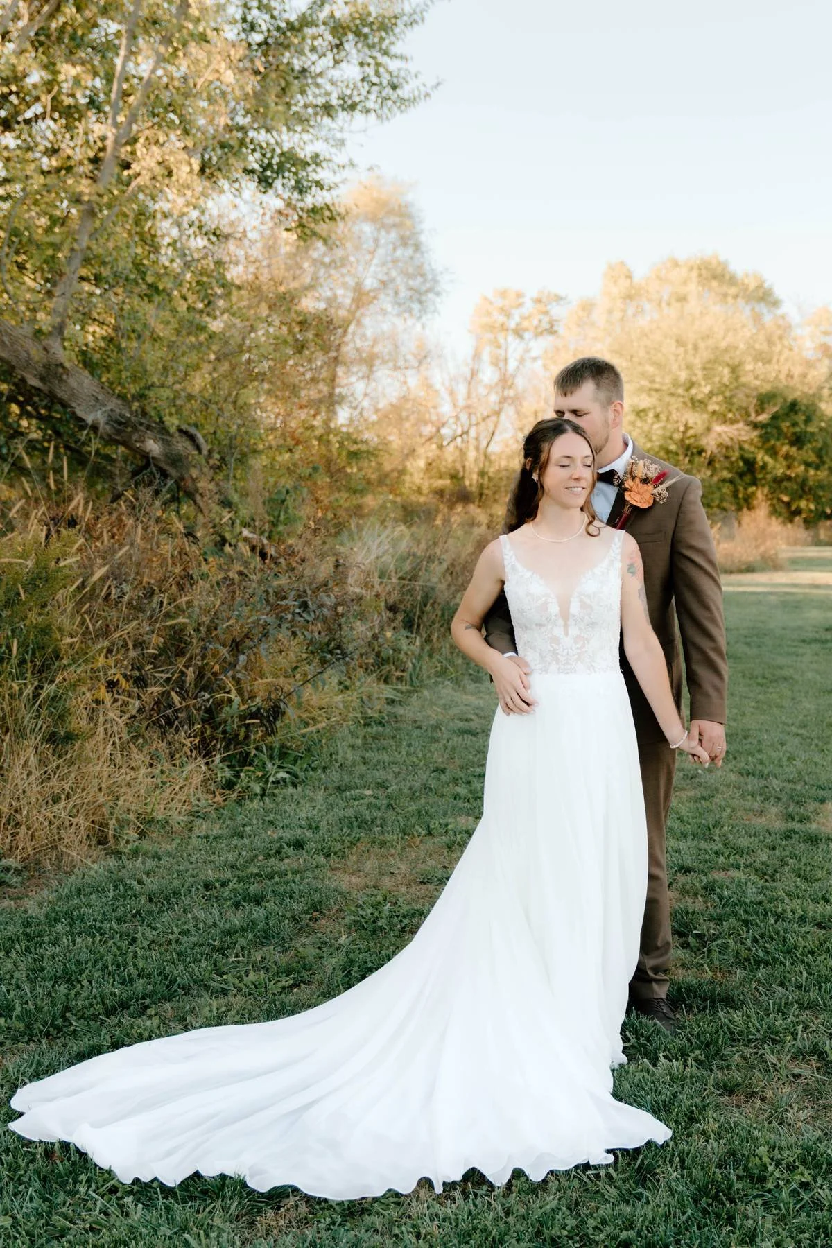 A bride and groom standing together outdoors on a grassy field during sunset, with the groom embracing the bride from behind and holding her hand while she smiles with eyes closed. The bride is wearing a white wedding dress with lace bodice and a lon