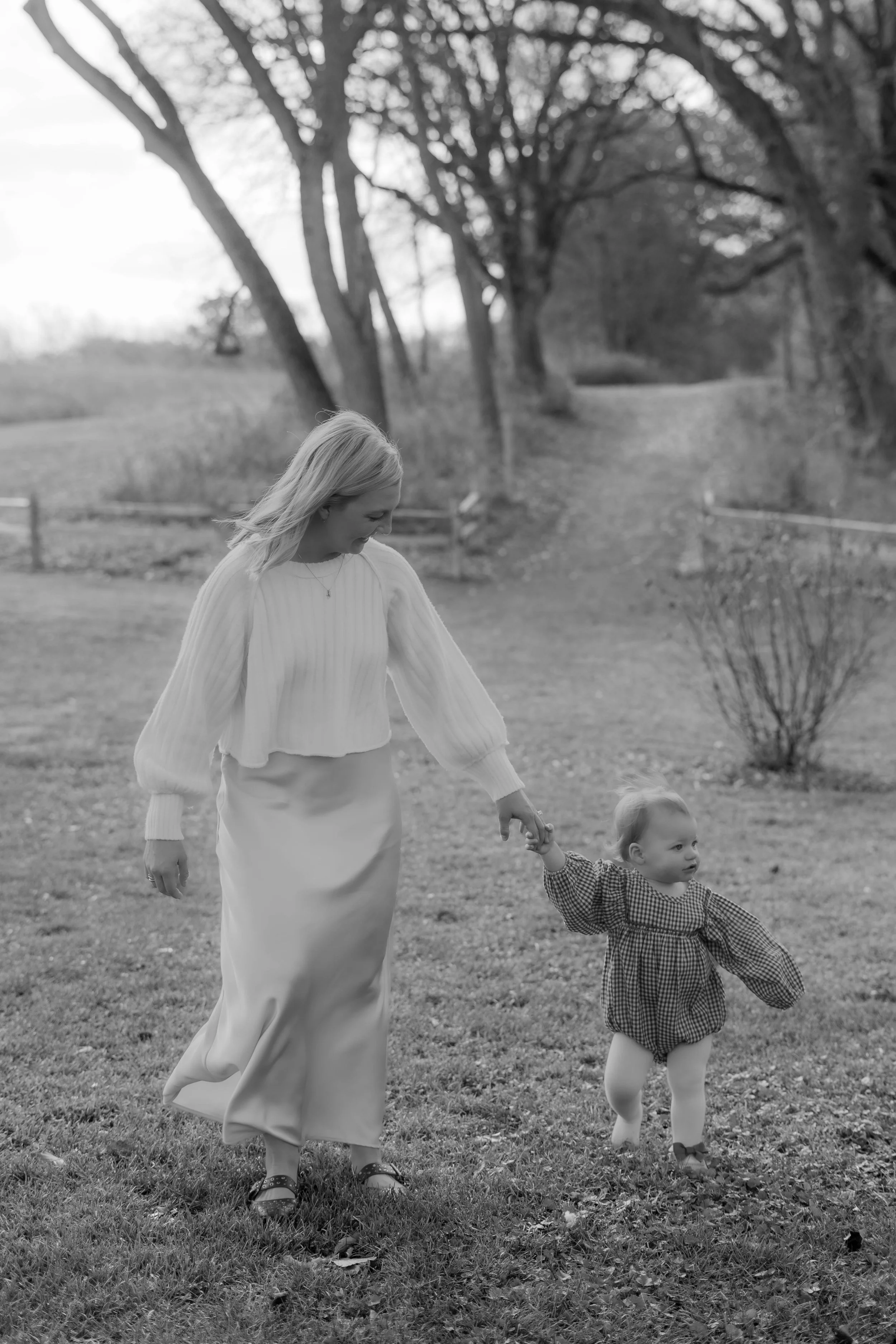 A woman in a long skirt and sweater holding hands with a young girl in a checkered dress outside in a park-like setting during the daytime.