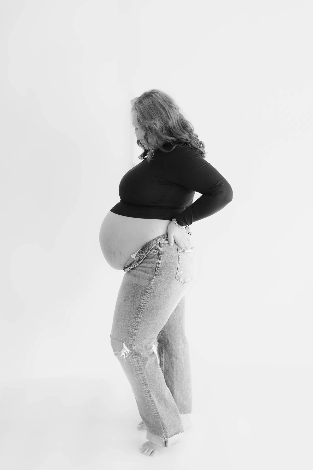 Black and white photo of a pregnant woman standing in profile, wearing a black long-sleeve top and ripped jeans, with her hand on her hip, against a plain light-colored background.
