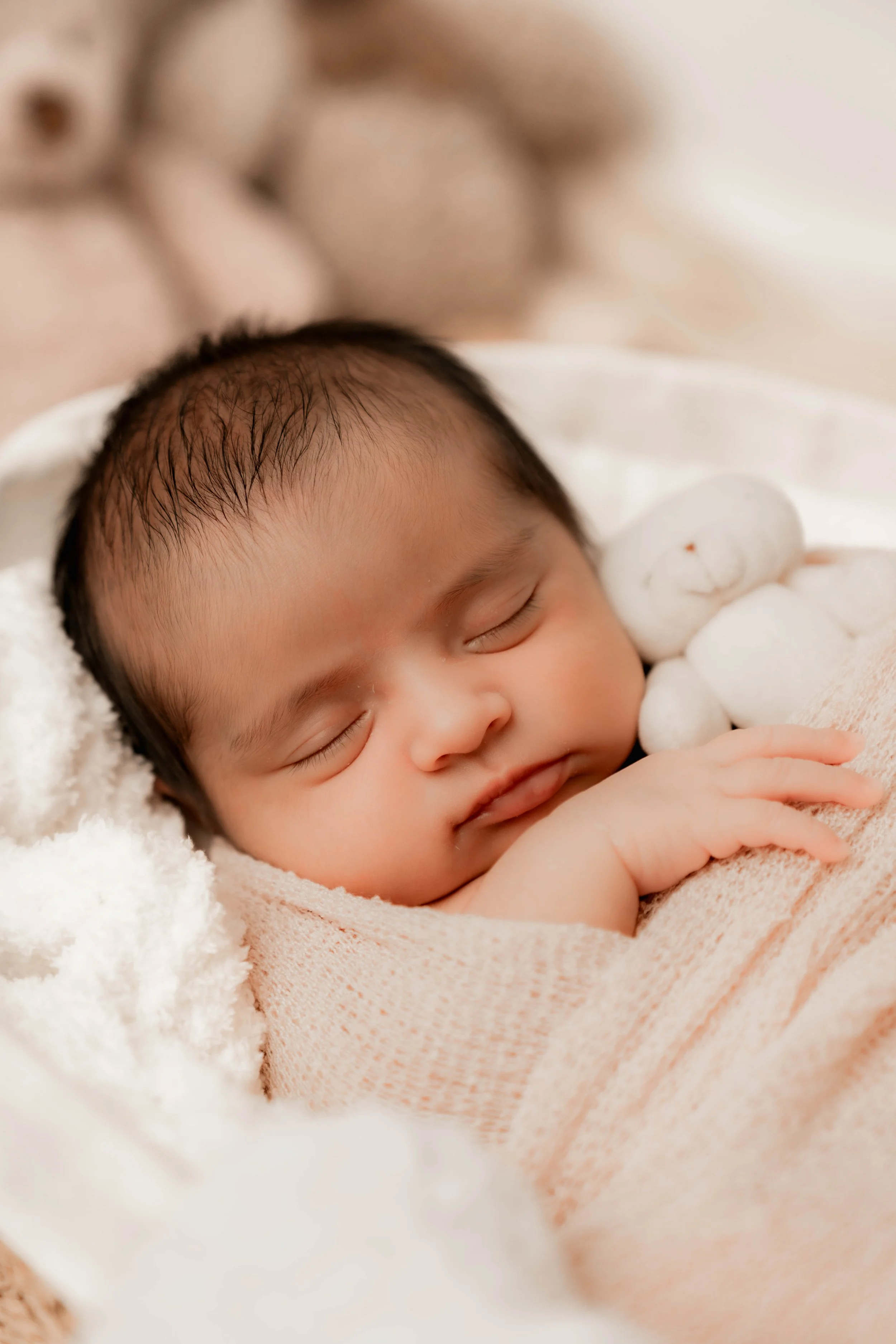 A sleeping baby cuddling a small white teddy bear, wrapped in a peach-colored blanket, with soft blanket textures surrounding them.