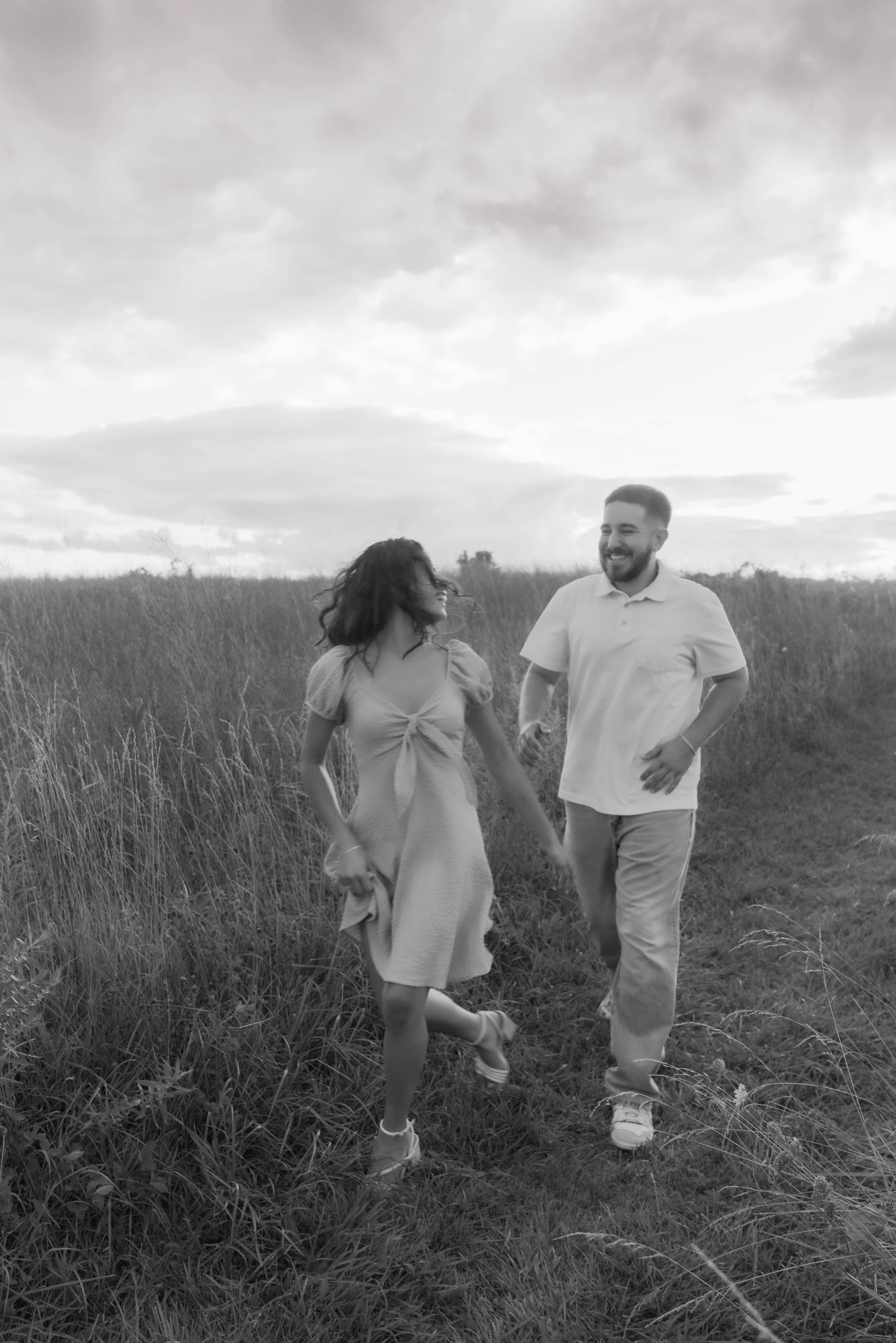 A black and white photo of a smiling couple holding hands and running through a grassy field outdoors with cloudy sky.