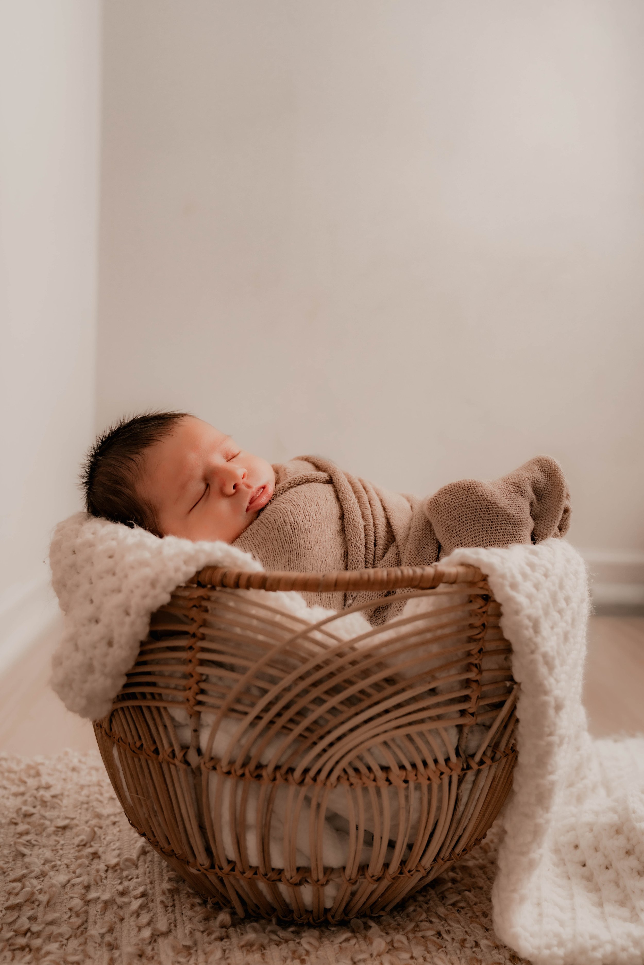 A sleeping baby wrapped in beige clothing, lying in a wicker basket with a soft blanket, on a textured beige rug against a plain light-colored wall.