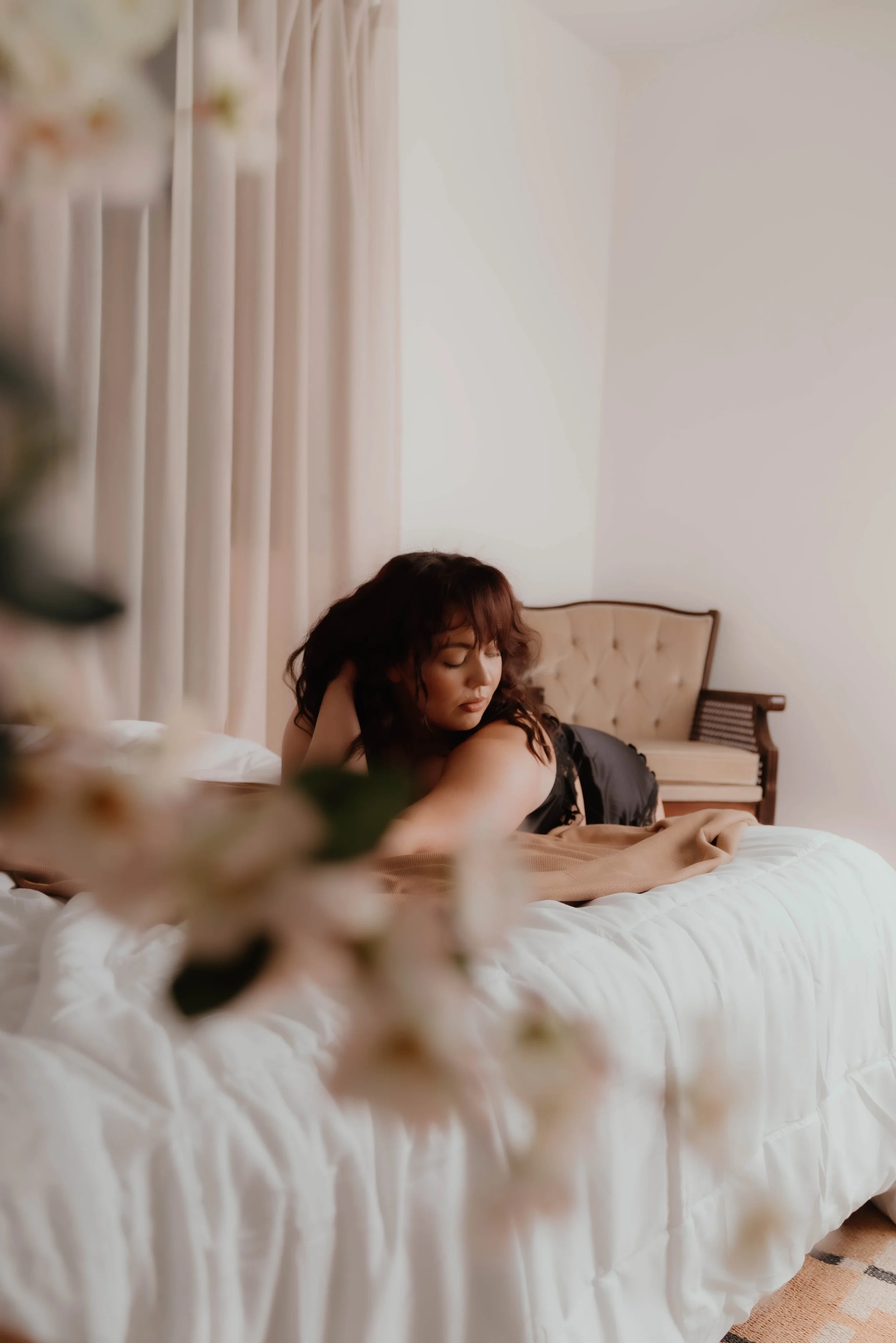 A woman with curly hair lying on her stomach on a bed, with a cream-colored headboard and light-colored curtains in the background.