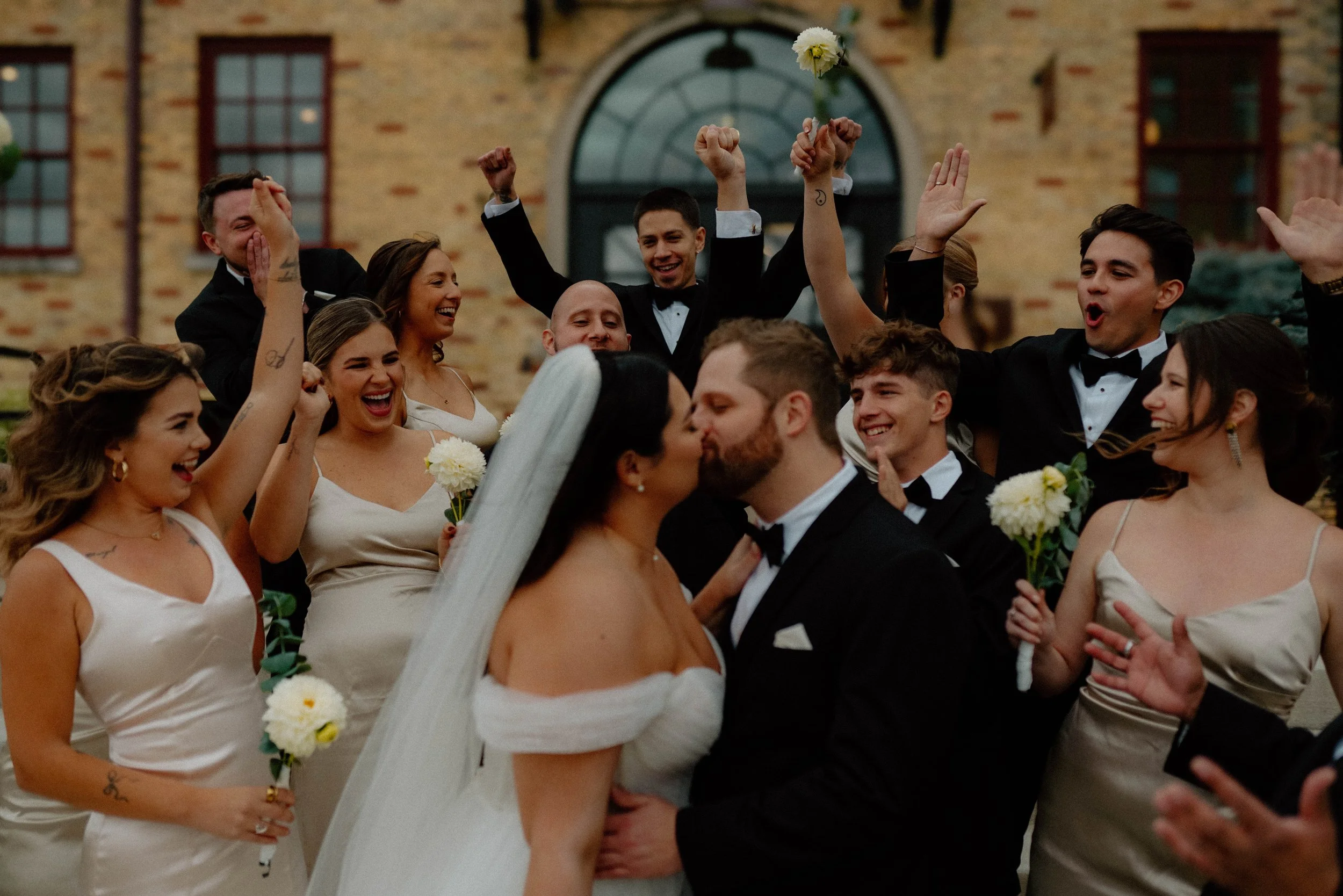 A group of people in wedding attire celebrating outdoors, with two people kissing in the foreground and others smiling and raising their hands in the background.