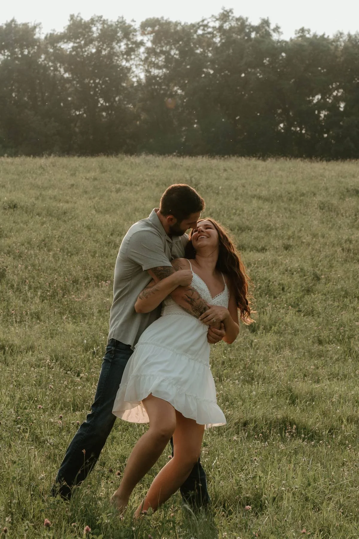 A couple dancing and smiling in a grassy field during sunset, surrounded by trees.