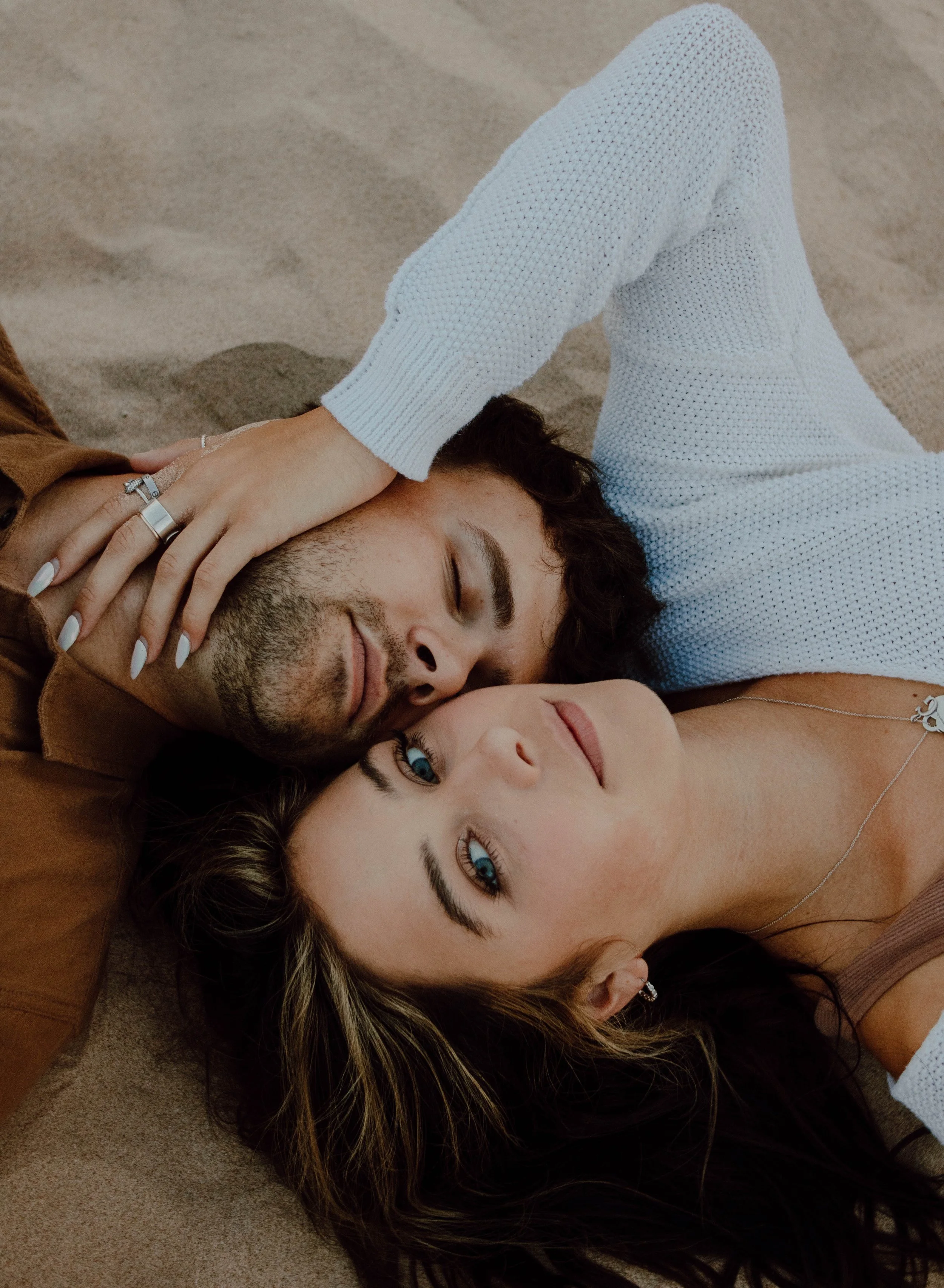 A young man and woman lying on the sand, close together, with the woman resting her head on the man, both gazing at the camera.