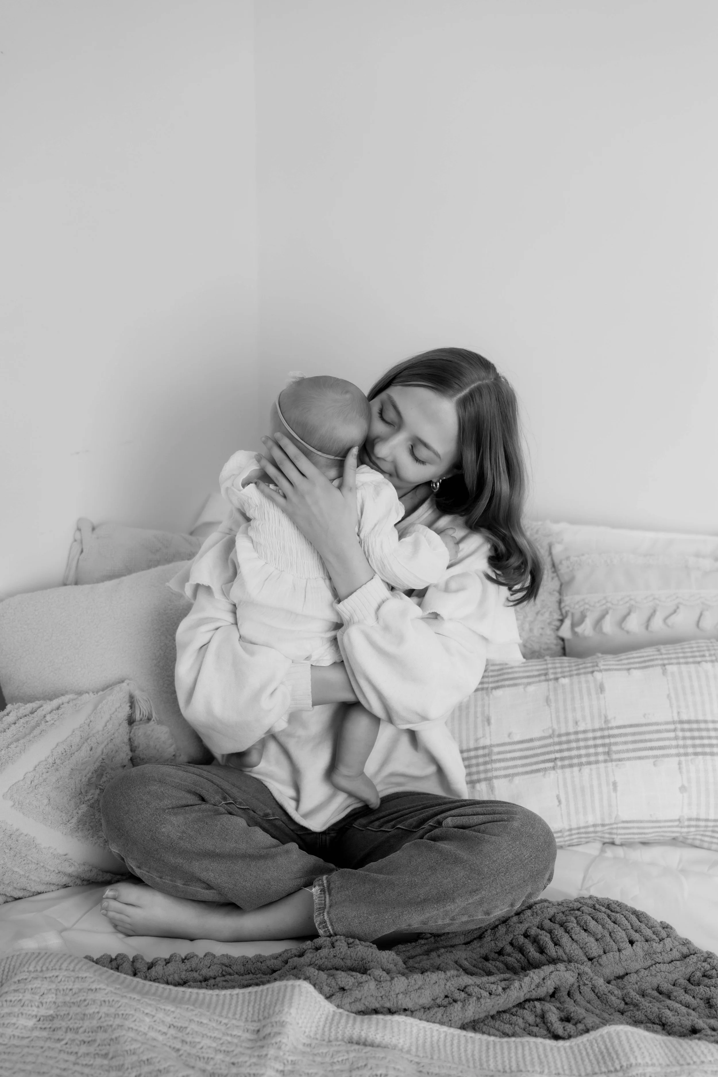 A woman sitting cross-legged on a bed, holding and hugging a young child close to her face in a warm embrace.