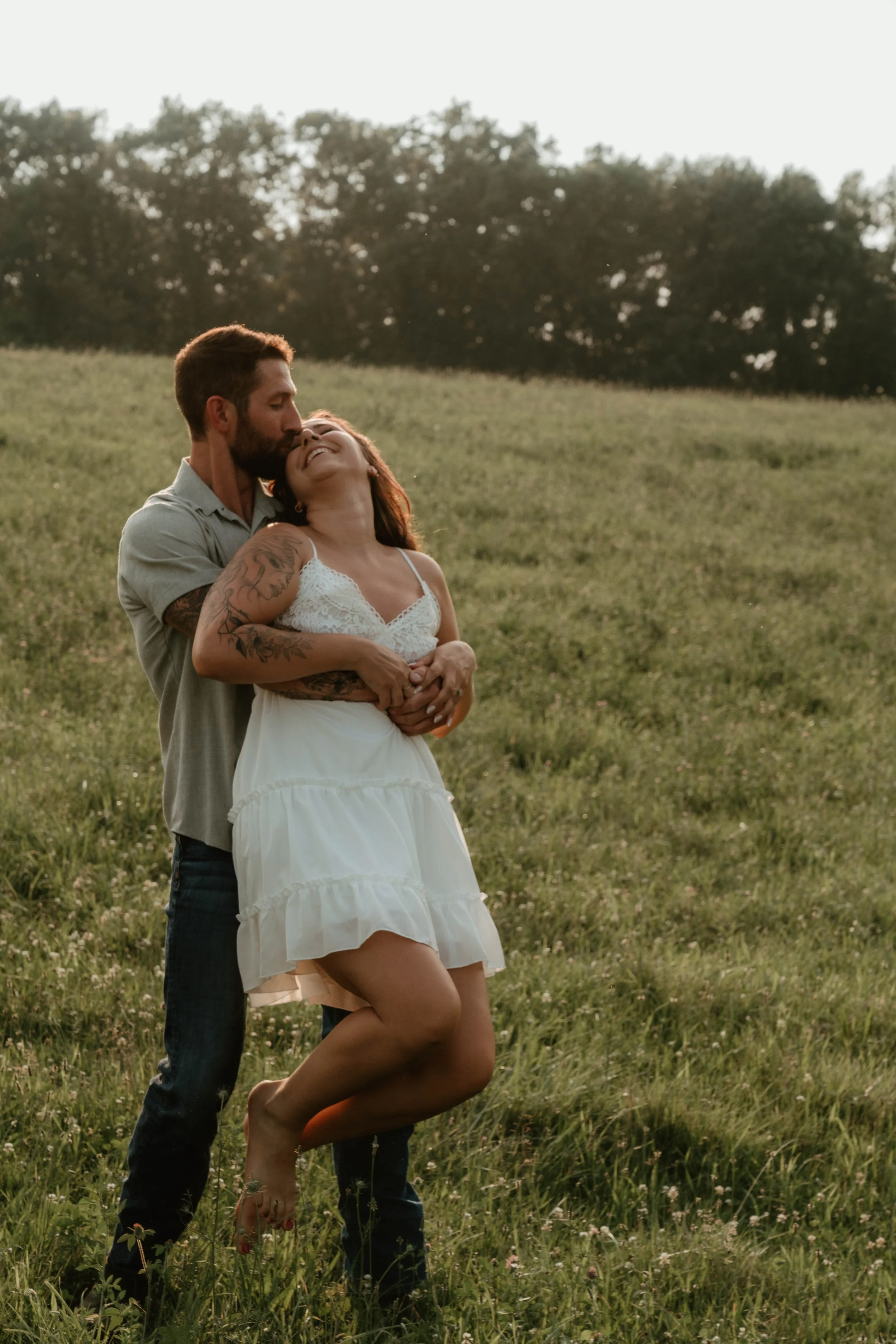 A man and woman embrace in a field during sunset, with trees in the background. The woman is smiling with her eyes closed, wearing a white dress. The man is kissing her on the forehead, dressed in a light-colored shirt and dark pants.