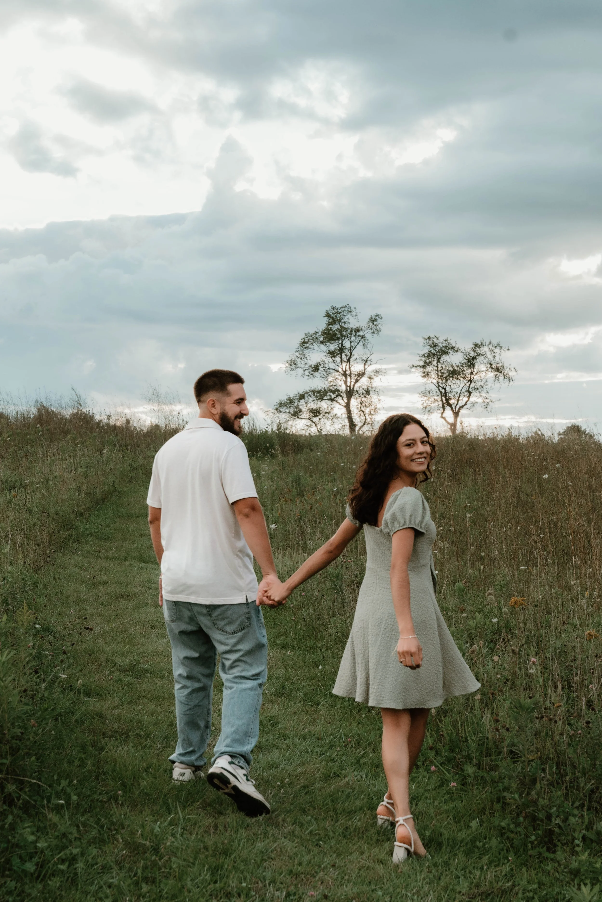 A young couple holding hands and walking on a grassy path in a field, with trees and cloudy sky in the background, smiling and looking back.