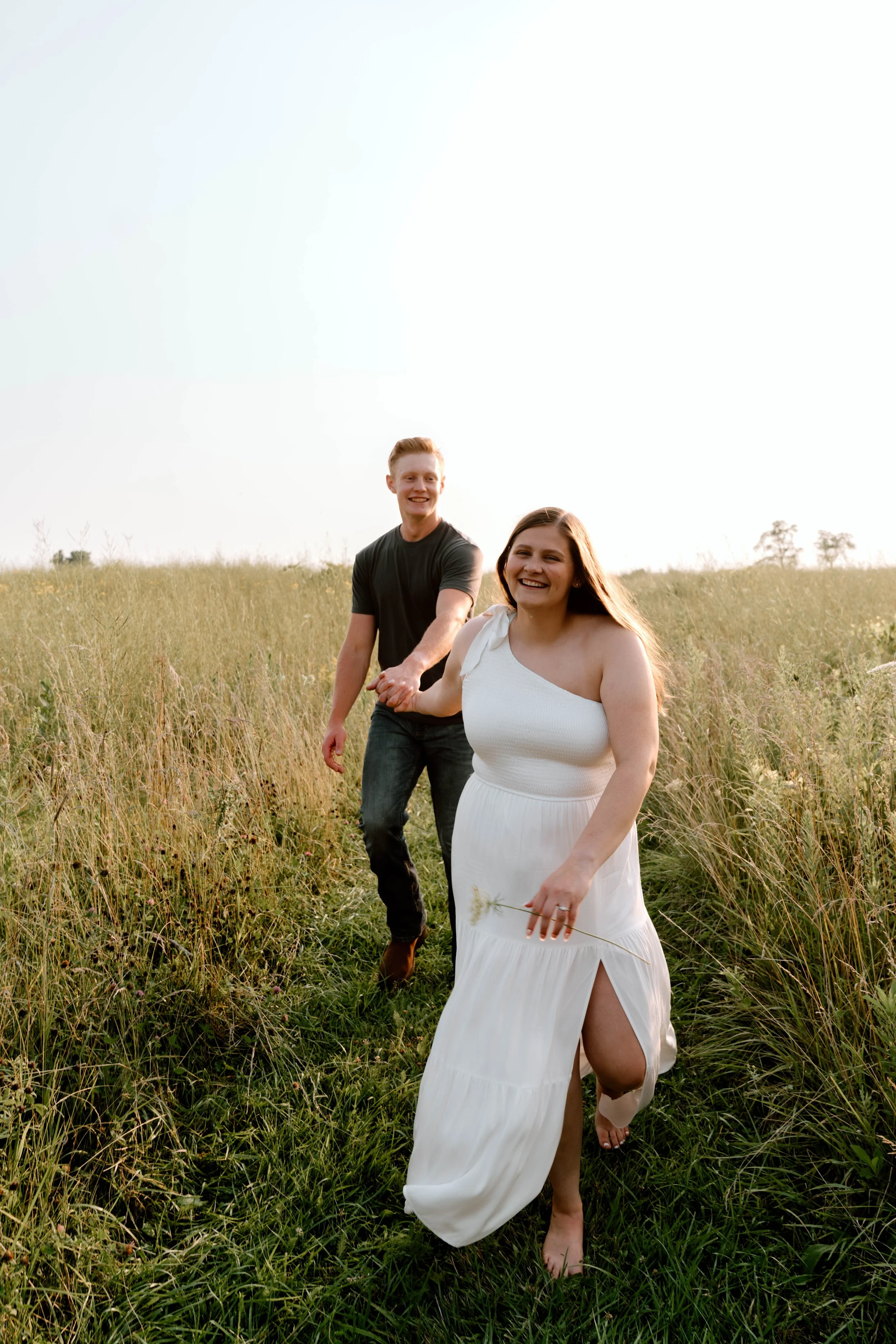 A couple holding hands and walking barefoot through a grassy field, smiling, with the woman in a white dress and the man in a black shirt.