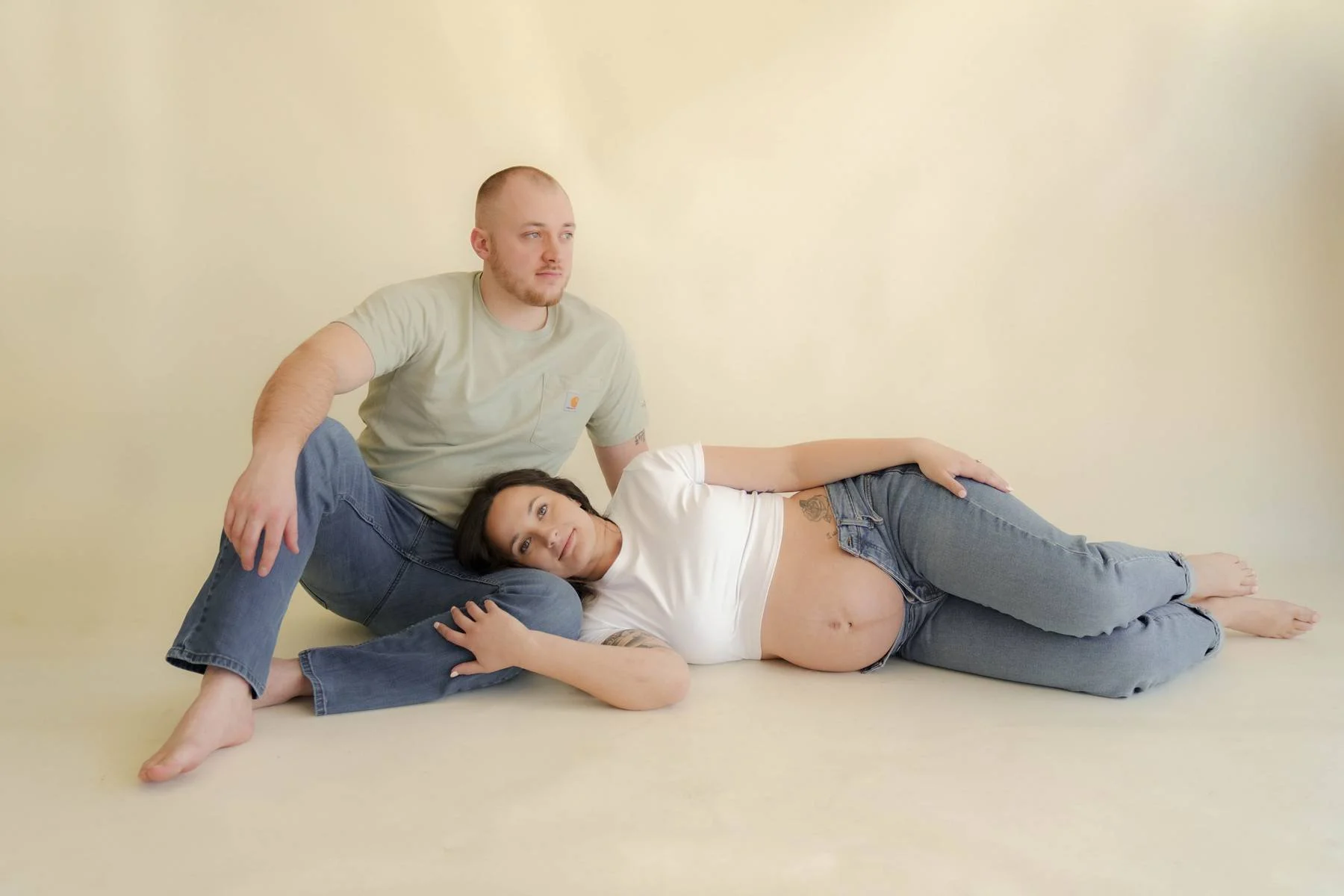 A couple with a pregnant woman lying on the floor and her partner sitting beside her, both casually dressed in jeans and t-shirts, against a plain light yellow background.