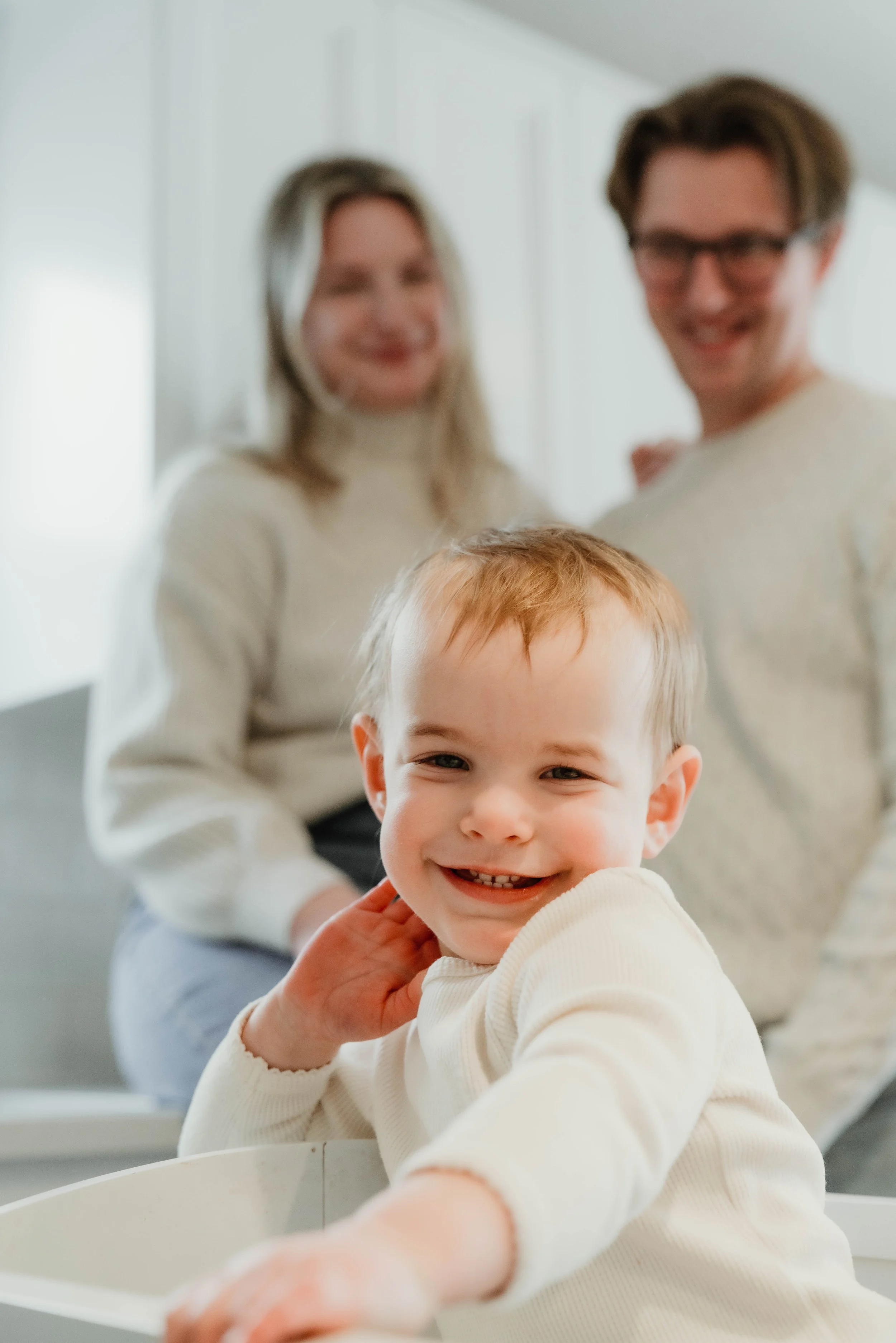 Young boy smiling and reaching out towards the camera, with a blurred family of two adults in the background indoors.