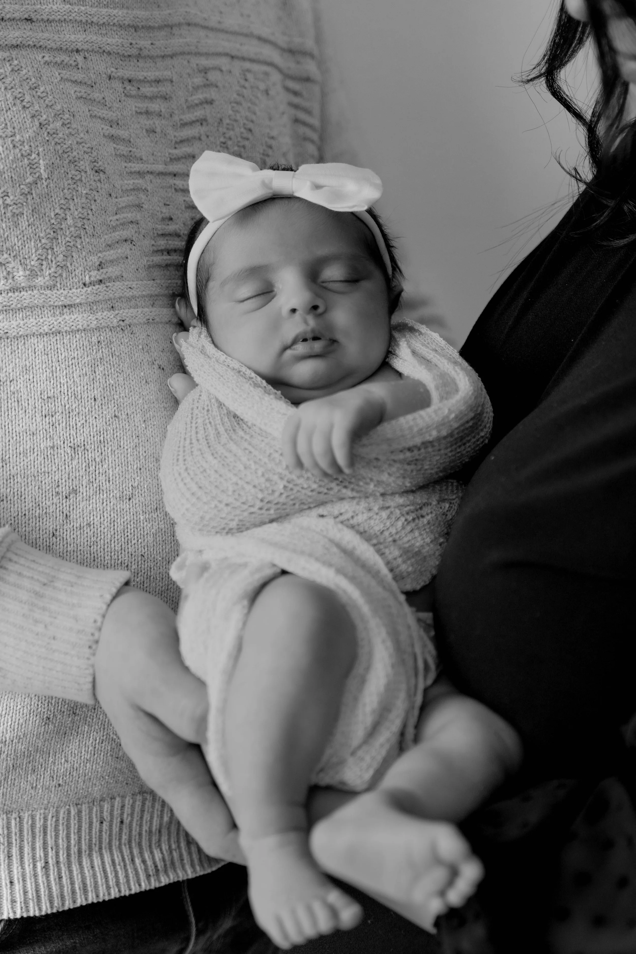 Black and white photo of a sleeping baby girl with a bow headband, wrapped in a knitted blanket, held by an adult wearing a dark top.