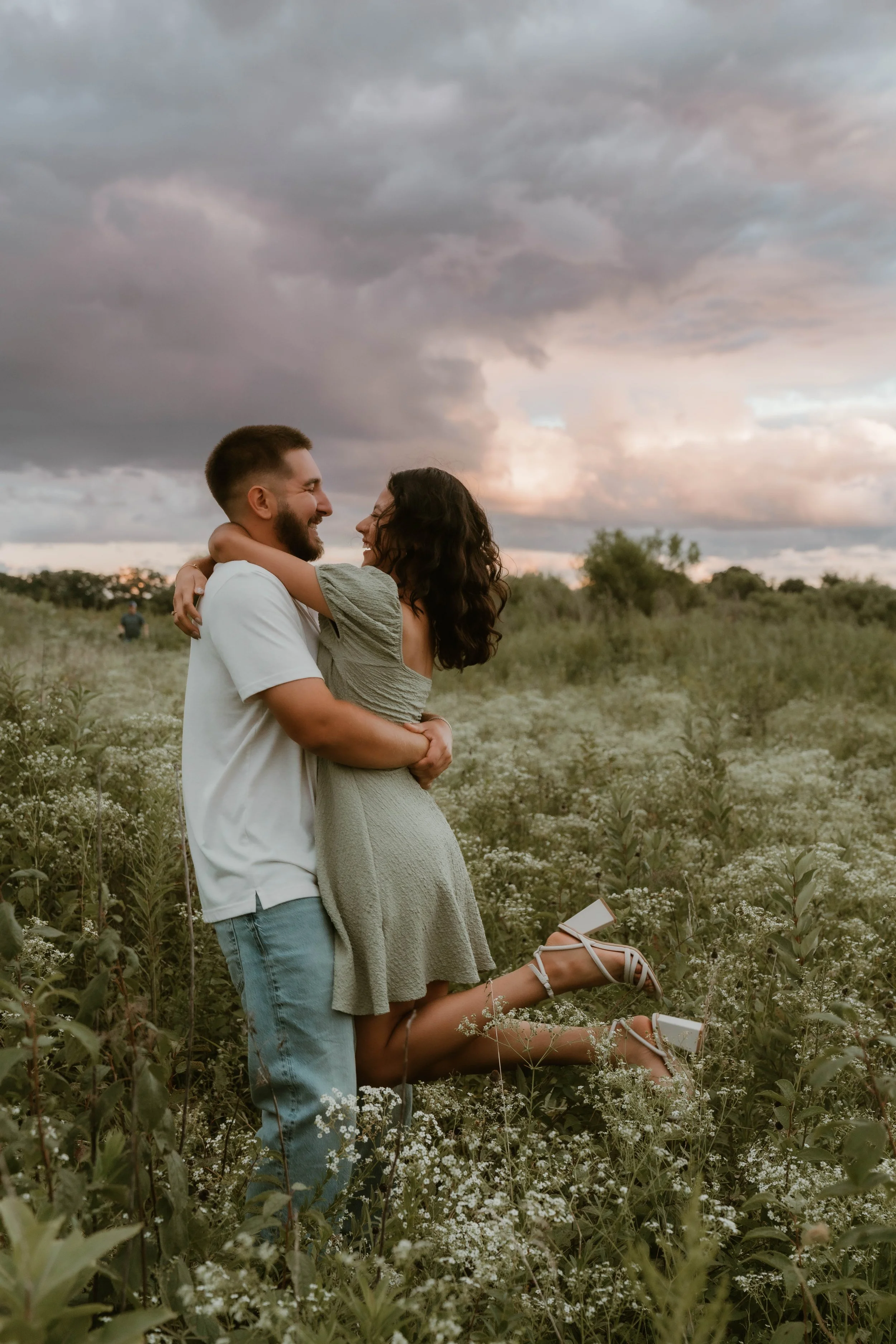 A happy couple embracing in a field of wildflowers under a cloudy sky, with the woman lifting the man, both smiling.