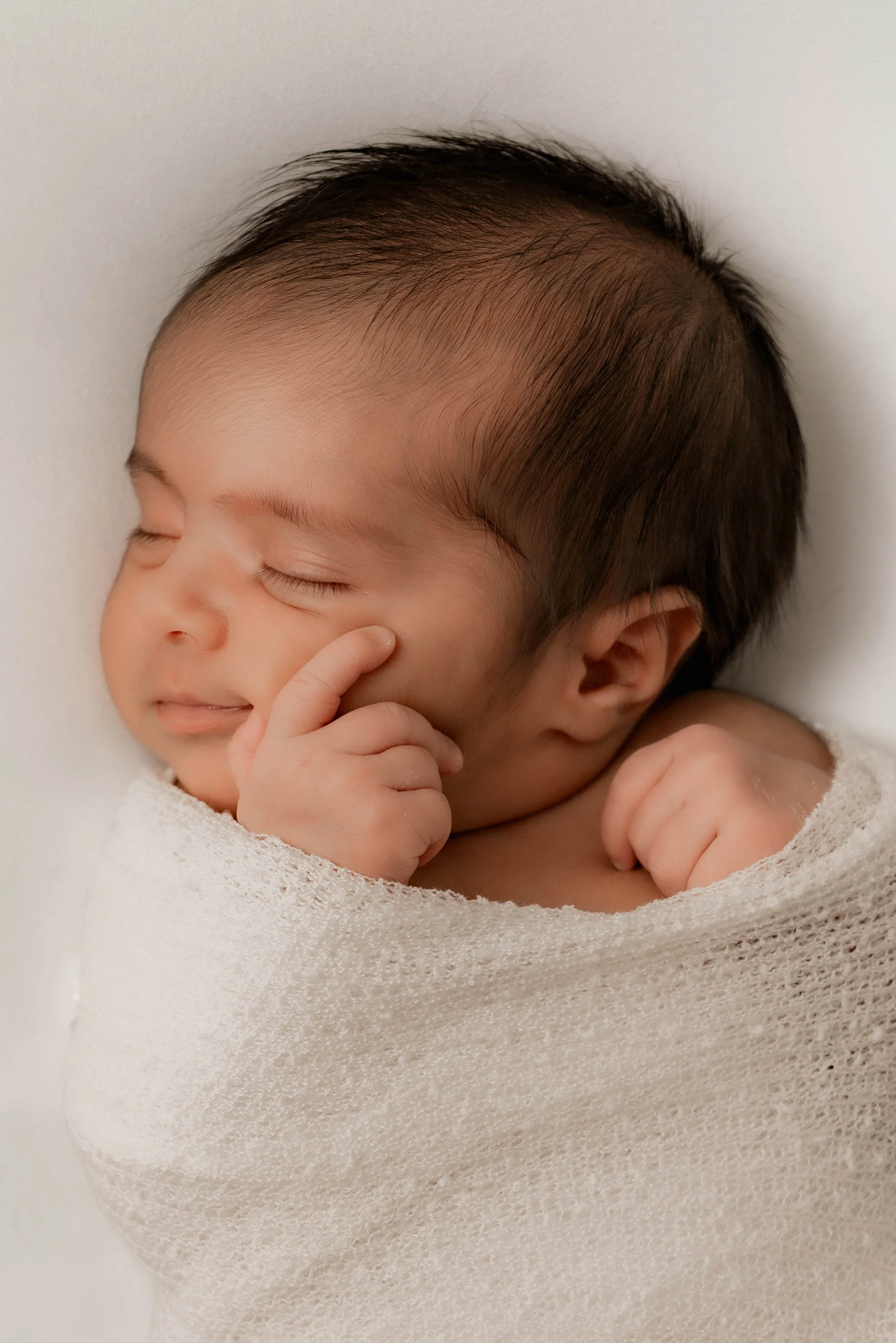 A sleeping baby wrapped in a knit blanket, resting peacefully with one hand near the face and a slight smile.