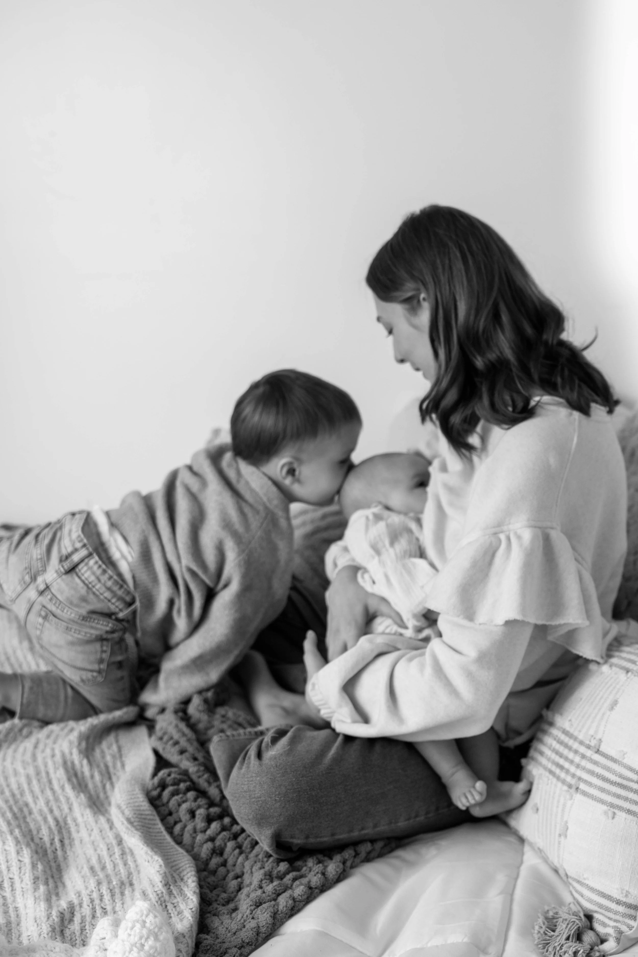 A woman sits on a bed holding a baby, while an older child leans over to kiss the baby's head, in a cozy indoor setting.